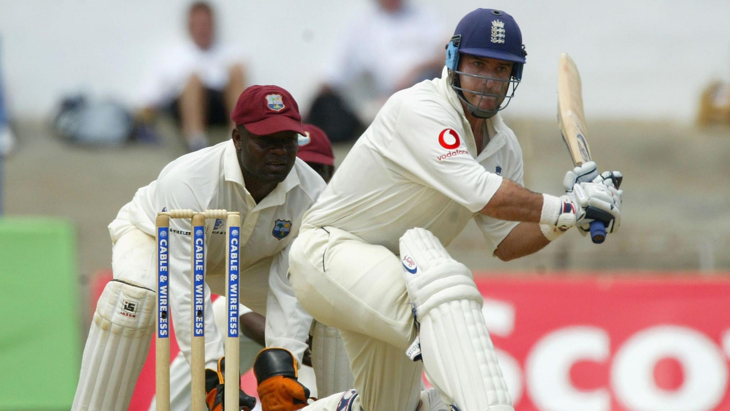 A man wearing all white swinging a cricket bat, while another man wearing gloves stands close by.