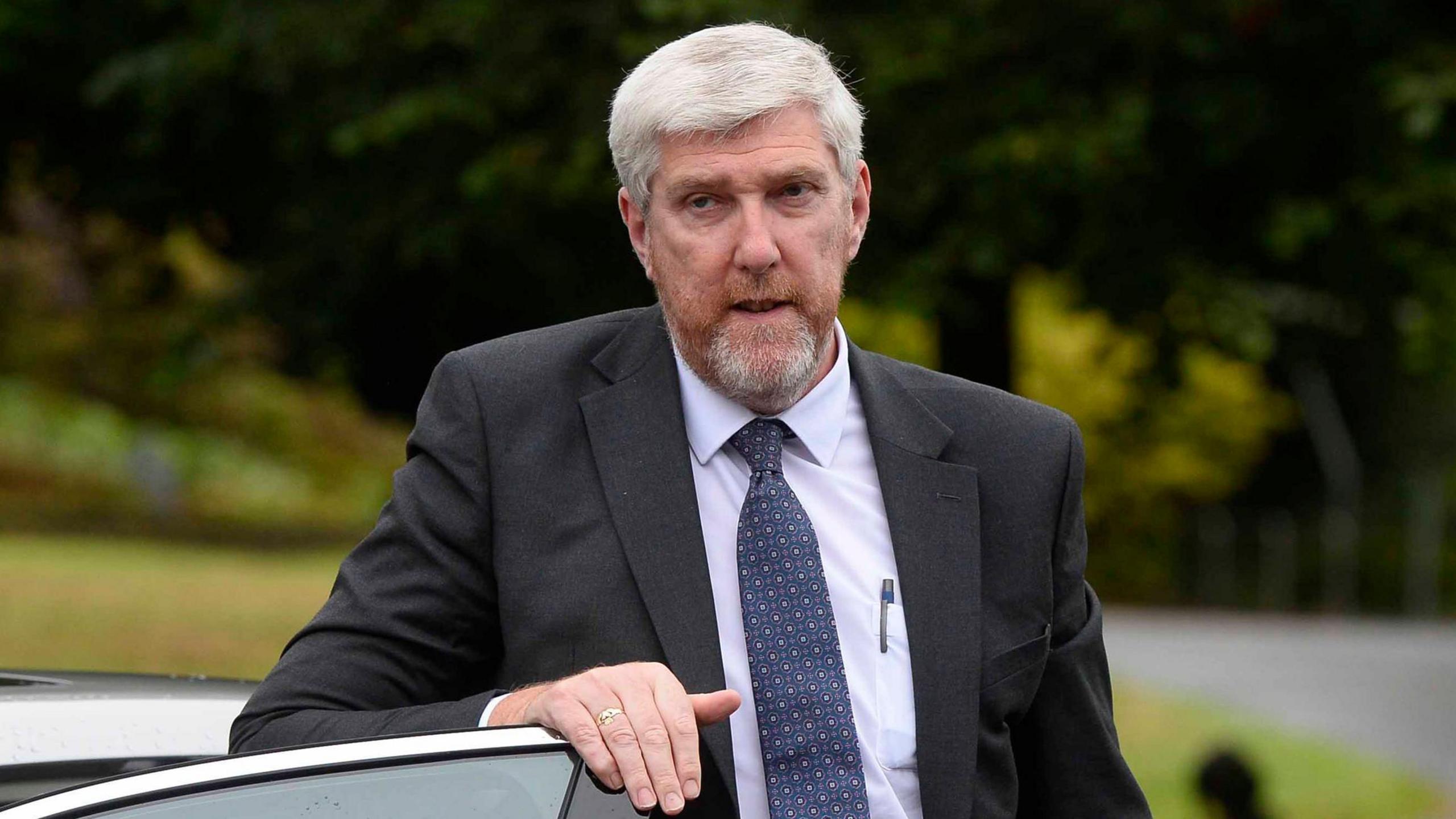 John O'Dowd - A grey-haired man wearing a dark suit jacket, a light-coloured collared shirt and a pattern tie. He is resting his hand on top of a car door with blurred shrubbery in the background.