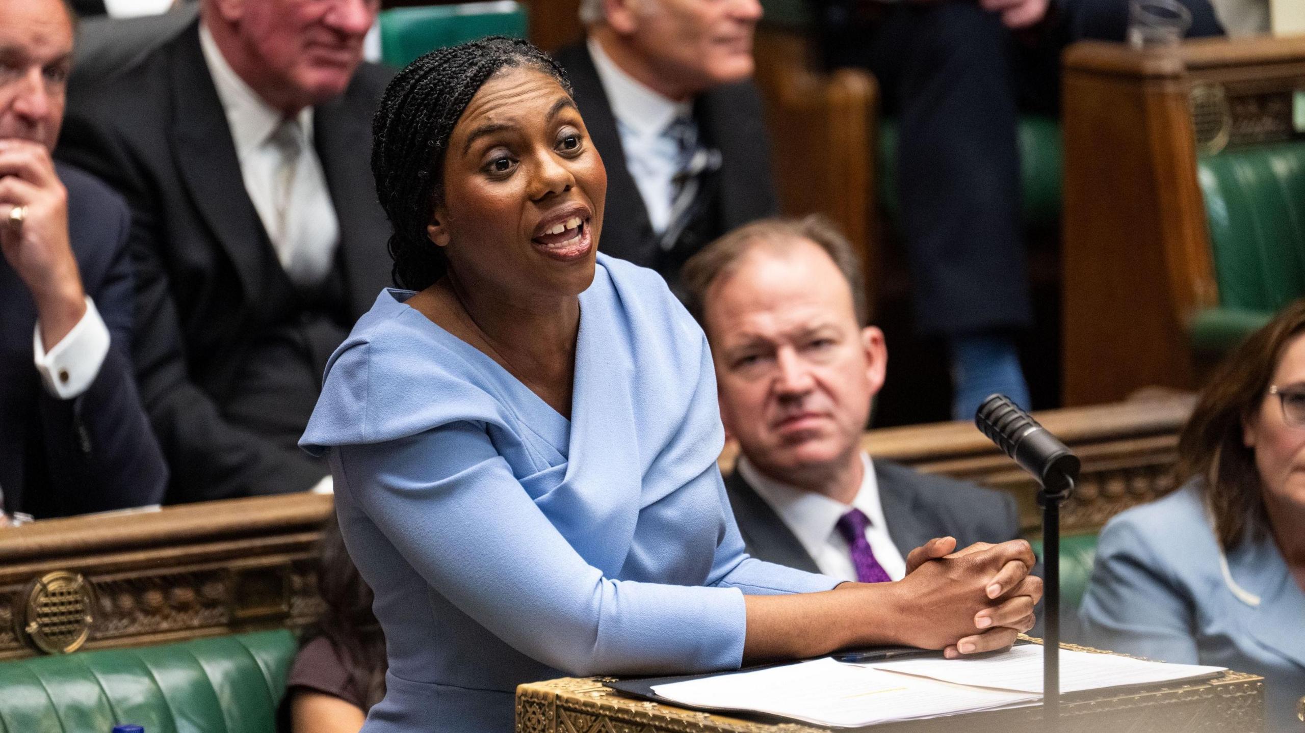 Kemi Badenoch stands in parliament addressing MPs. She wears her hair in long braid and is wearing a sky blue dress.