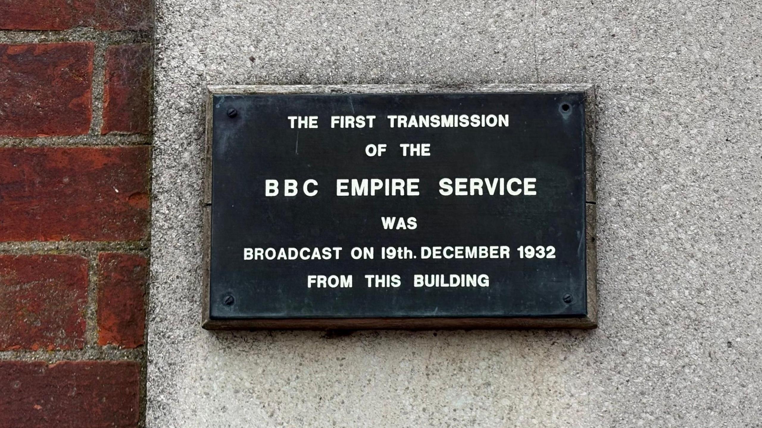 A plaque with white lettering on a black background. It says: "The first transmission of the BBC Empire Service was broadcast on 19th December 1932 from this building. The plaque is fixed to a wall of small stones and there is a brick wall to the left.