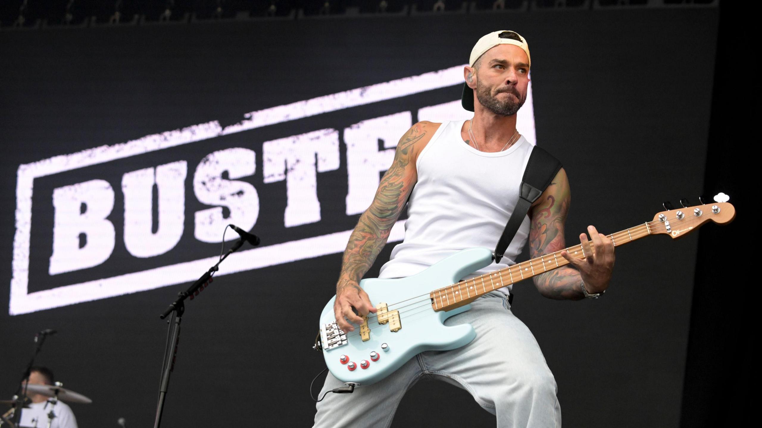 Busted's Matt Willis is on stage playing a very light blue bass guitar. He wears white jeans, a white vest and a white backwards cap. He stands behind a microphone, in front of a screen that reads "Busted" in white letters.