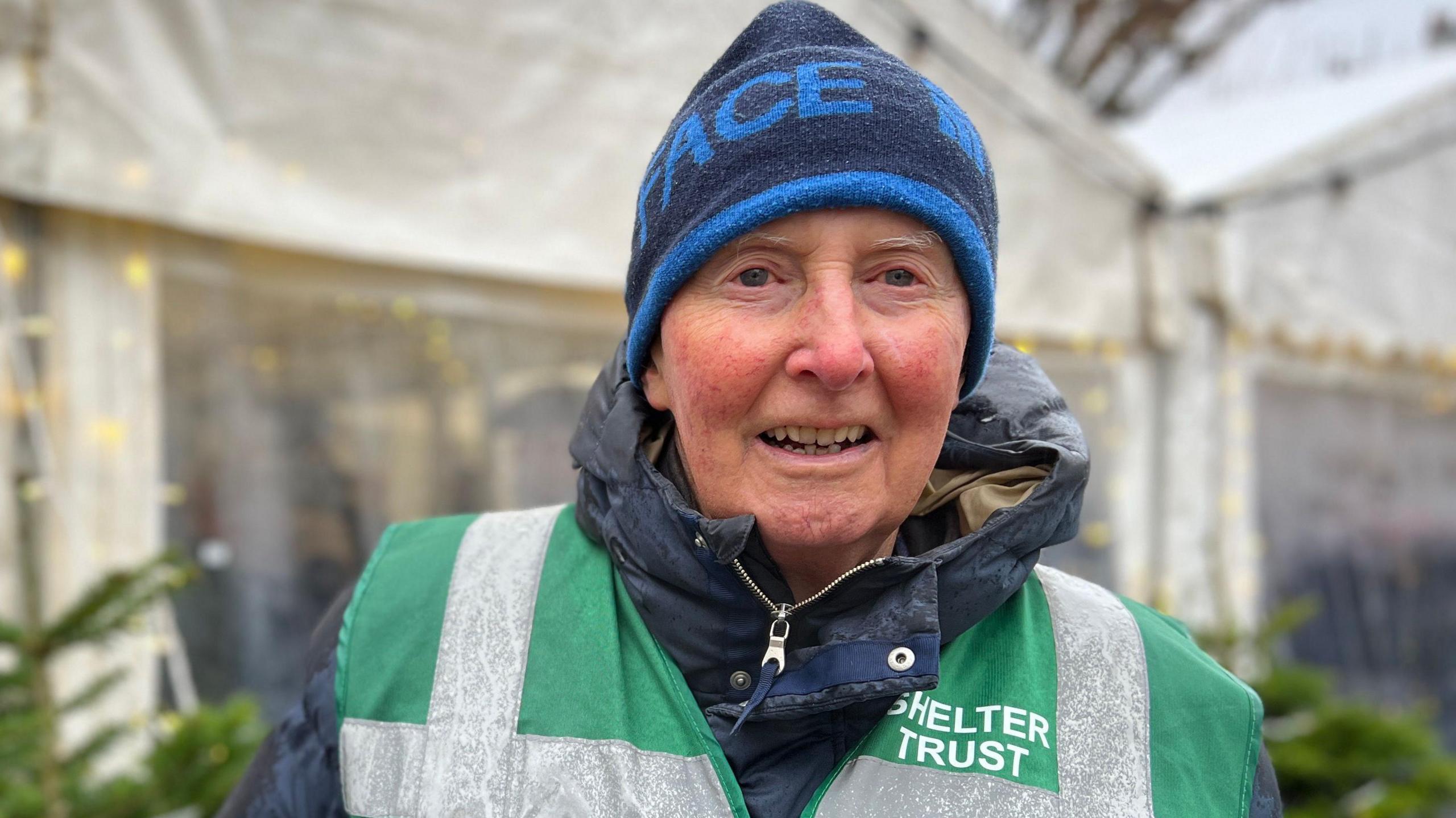 Richard Robins wears a dark blue and royal blue woolly hat with a navy blue coat and a dark green high viz jacket over the top that reads "Shelter Trust" on his left shoulder.