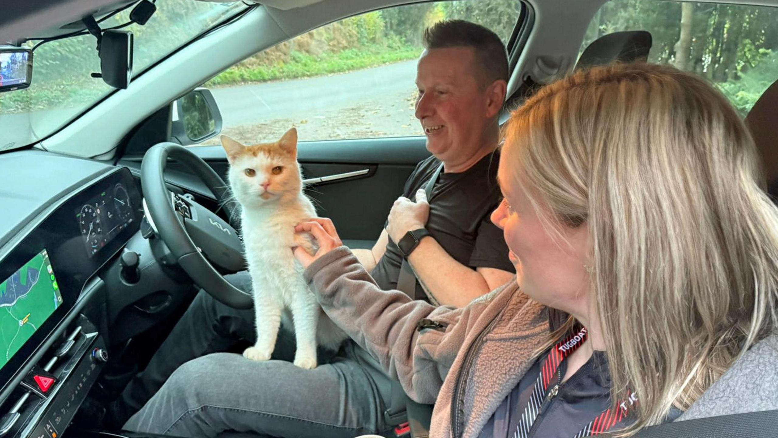 Beans the cat, white and ginger, sitting on the lap of Colin McBurnie, who is wearing a dark t-shirt and grey jeans. He is sitting in the driver's seat of a car. On the dashboard, a GPS screen is lit up in a green colour. Cara McBurnie is stroking Beans just above his front left leg. She is blonde and is wearing a grey hoodie with a red and black striped lanyard around her neck.