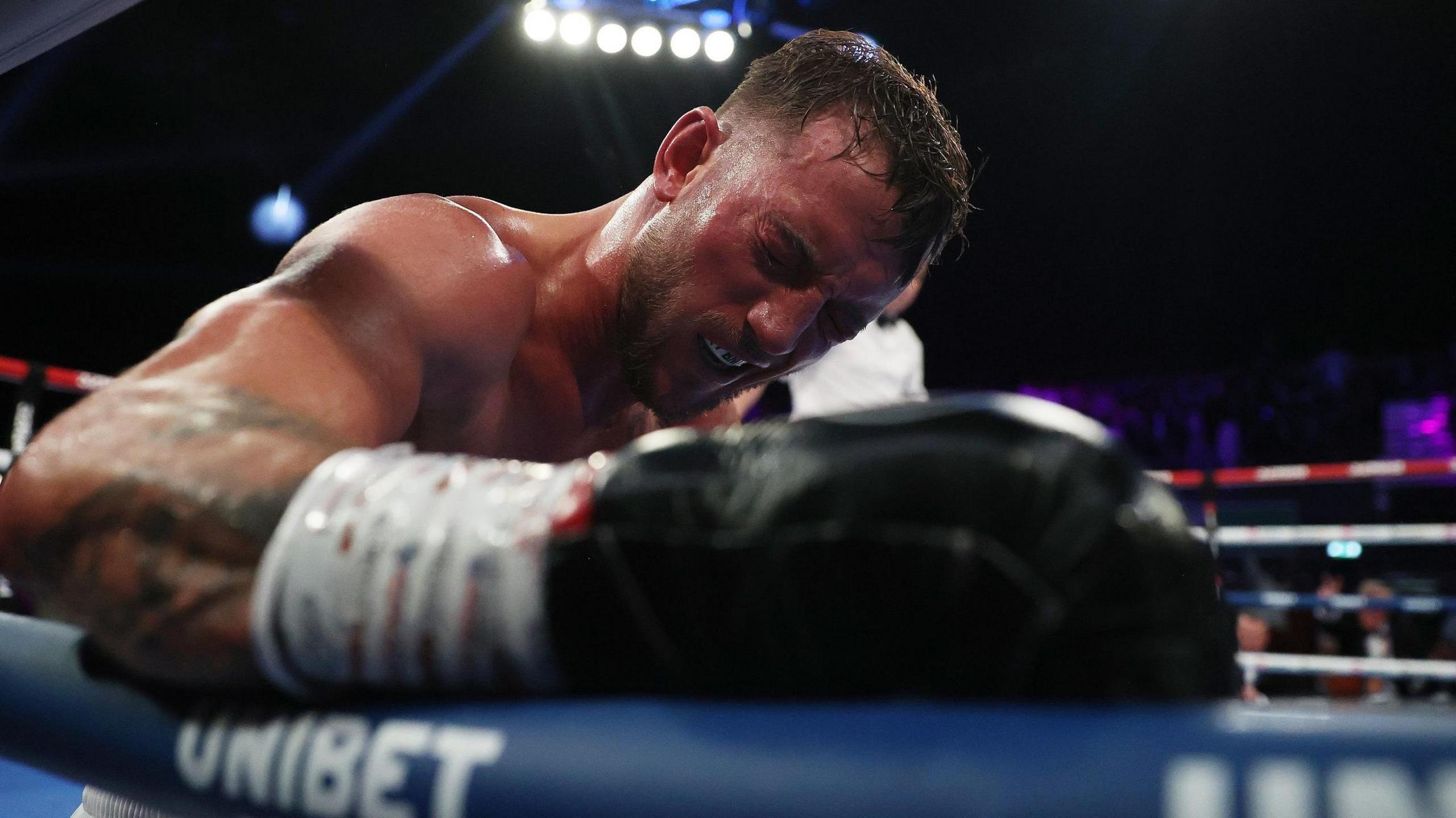 Brad Pauls pictured after being knocked down by Denzel Bentley. He is closing his eyes in pain and leaning on the ropes of the ring.