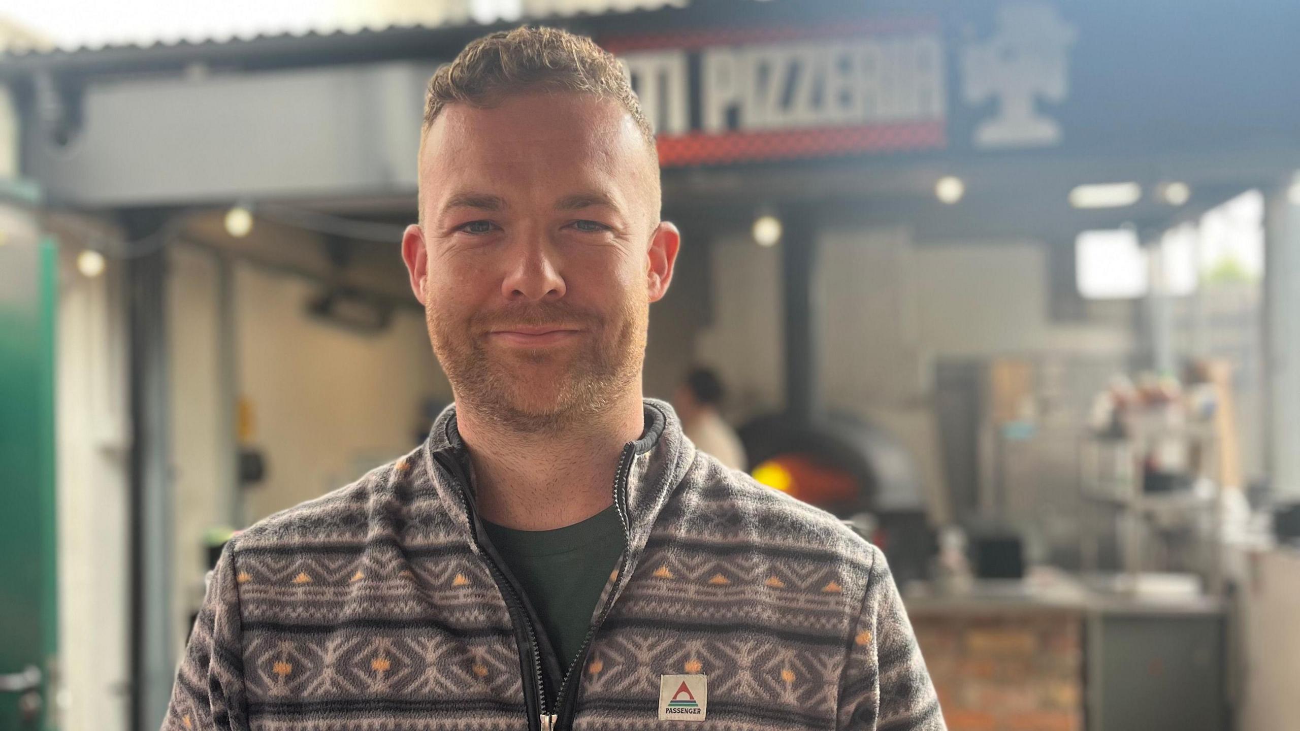 William Mayne, wearing a grey fleece, stands in front of the pizza preparation area outside his bar