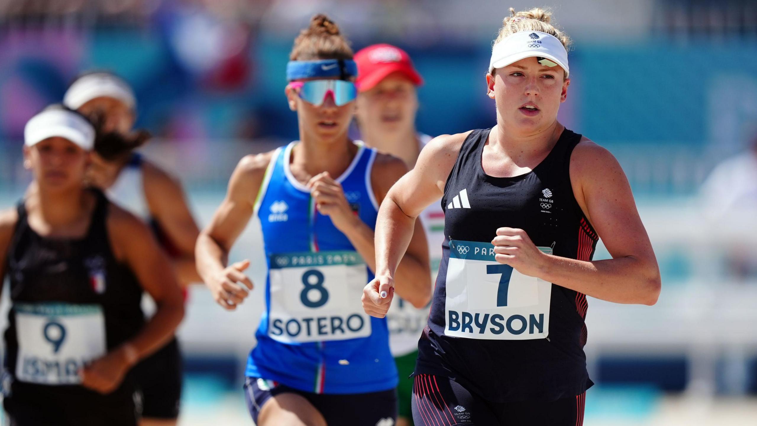 The image shows a running race with Kerenza in the foreground of the image in the lead. She is wearing a navy blue Team GB running vest and shorts with a white sun visor. She has her hair tied up and her running number on the front of her top which says '7 Bryson'. You can also see other competitors in the background who are also wearing running vests and shorts. They also have their numbers and names on the front of their tops. 