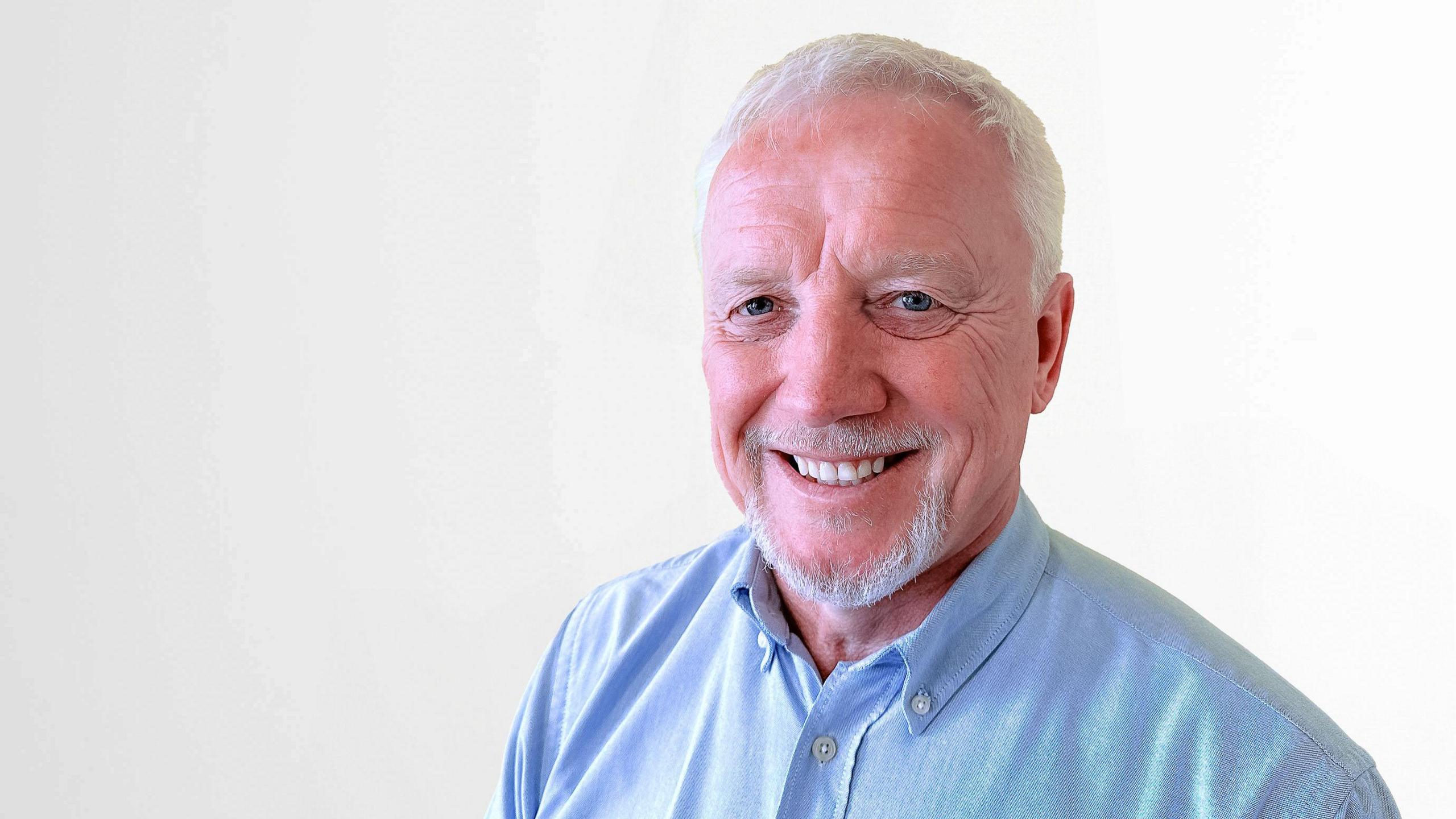 A man stood looking directly at the camera. He is dressed in a light blue button-up shirt and has a short white beard and white hair. The background behind him is plain white.