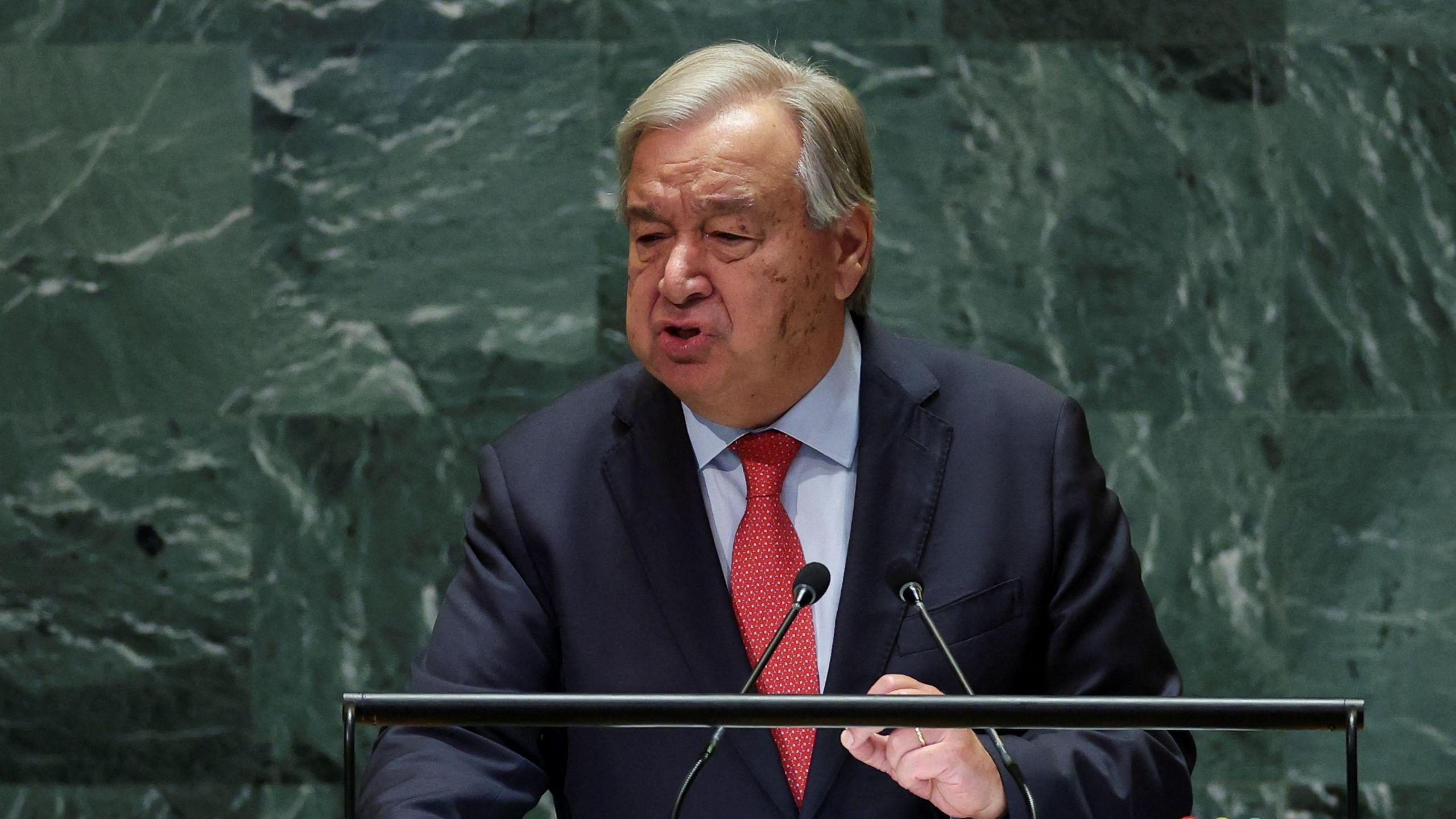 UN Secretary General Antonio Guterres giving a speech at the United Nations in New York. He is standing against the distinctive green marbled backdrop at a lectern, wearing a red tie, dark suit jacket and blue shirt