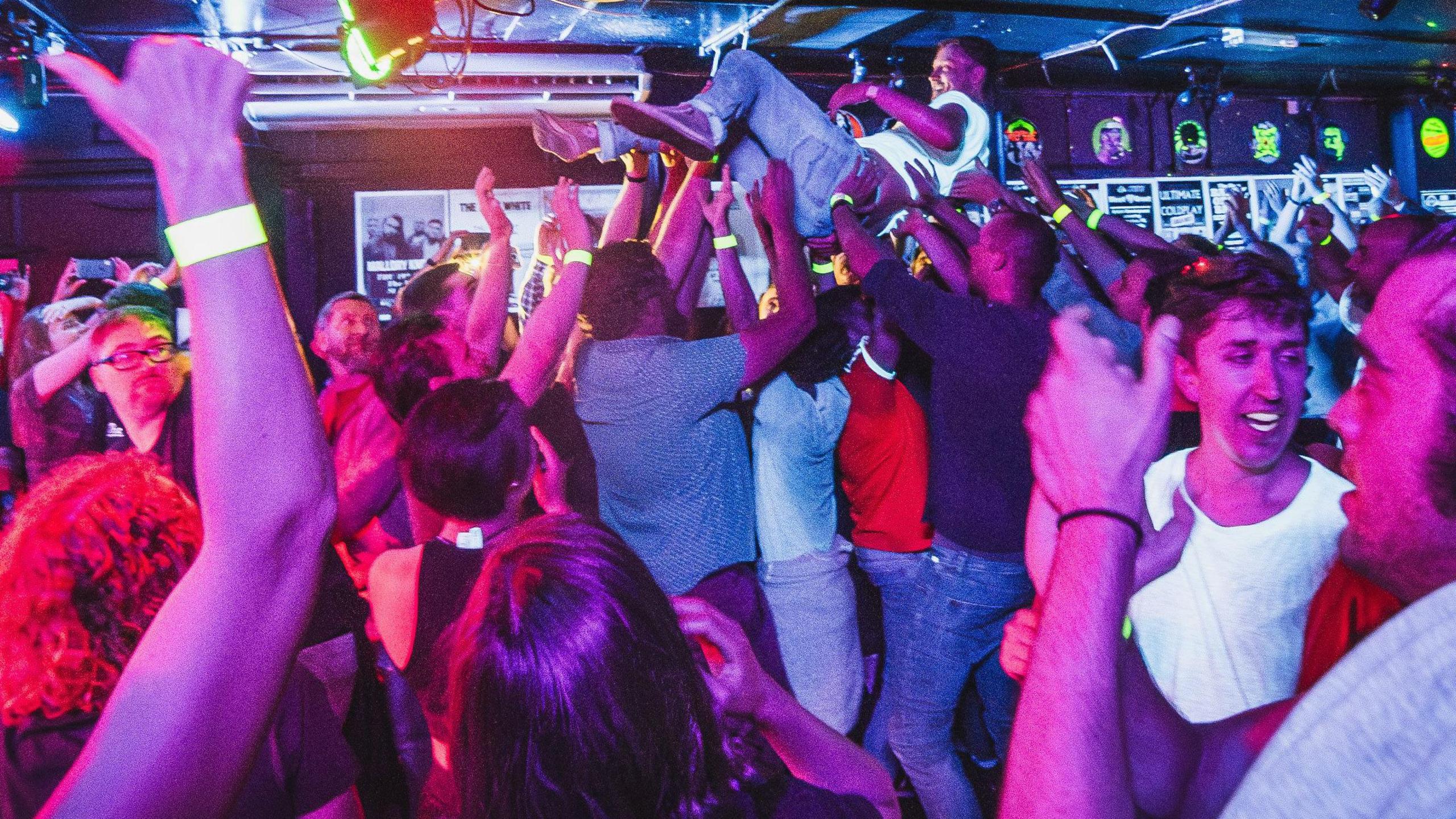 A room full of people in a music venue, one man has been lifted up, crowd surfing, while more people are standing around around waving their arms. 