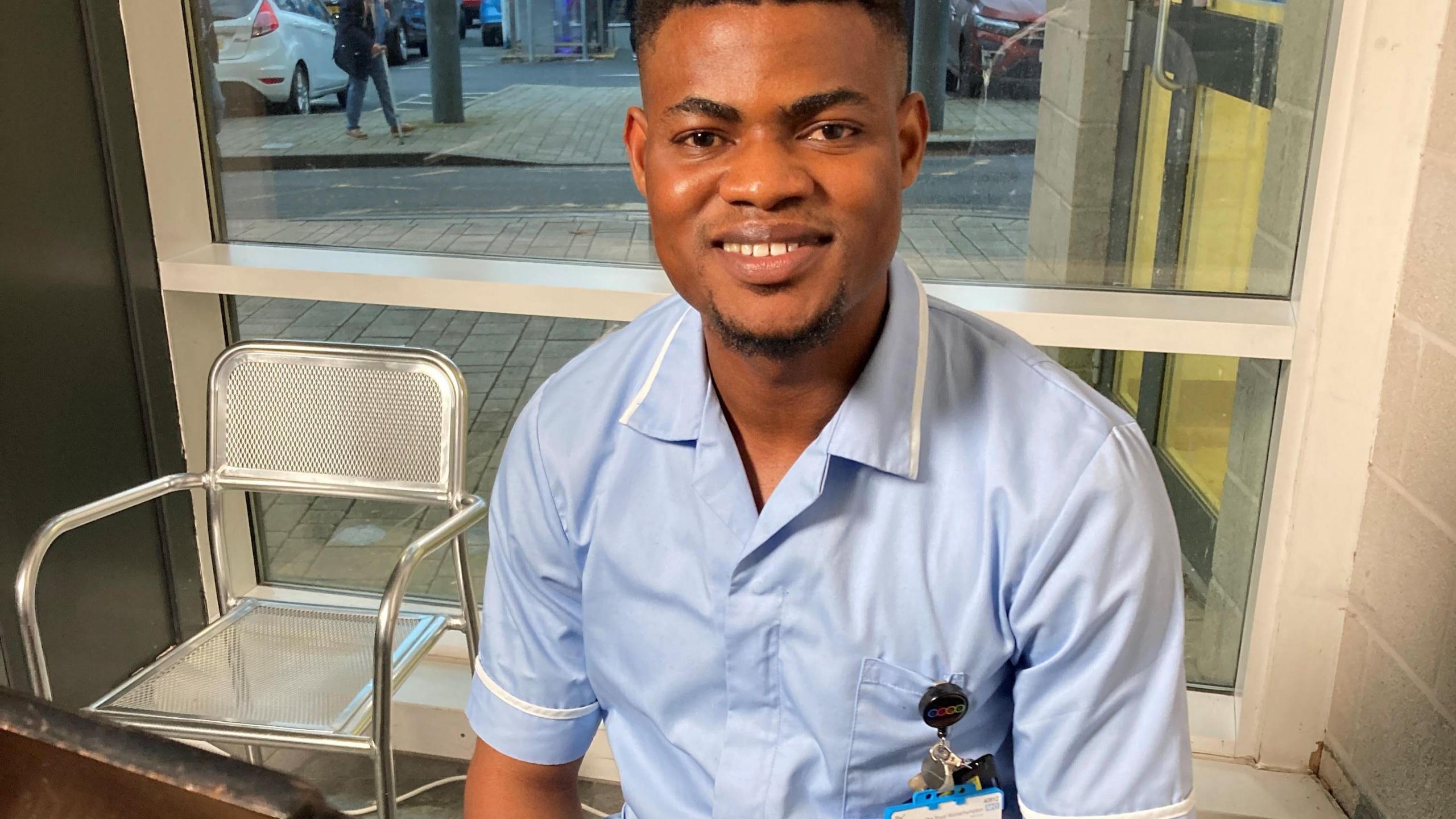 Daniel Aboyeji smiling at the camera. He is wearing a blue nursing uniform and has a work badge on. Behind him is a metal chair. 