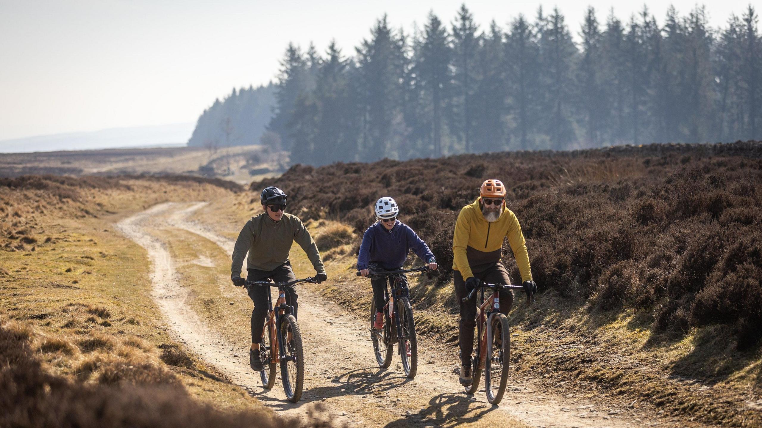 Three cyclists riding on a gravel route with some trees in the background. Two of the cyclists are men, one is wearing a green top and a black helmet, the other is wearing a mustard hoodie and an orange helmet. The other cyclist is a woman and is wearing a blue hoodie and a grey helmet.