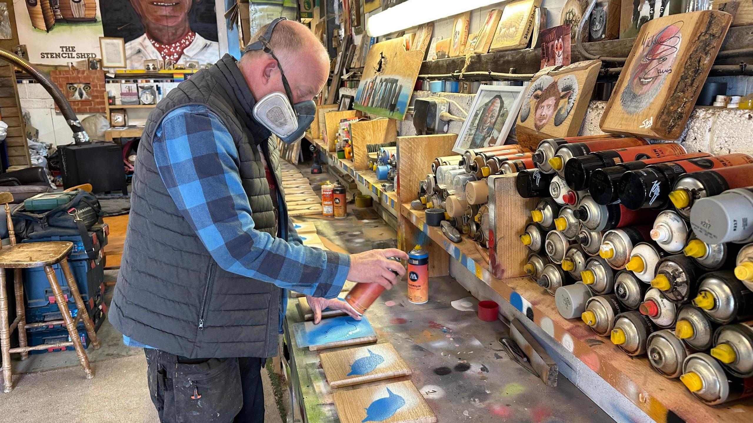 Syd wearing a mask and spray pairning some wooden blocked in his workshop, surrounded by spray cans and artwork