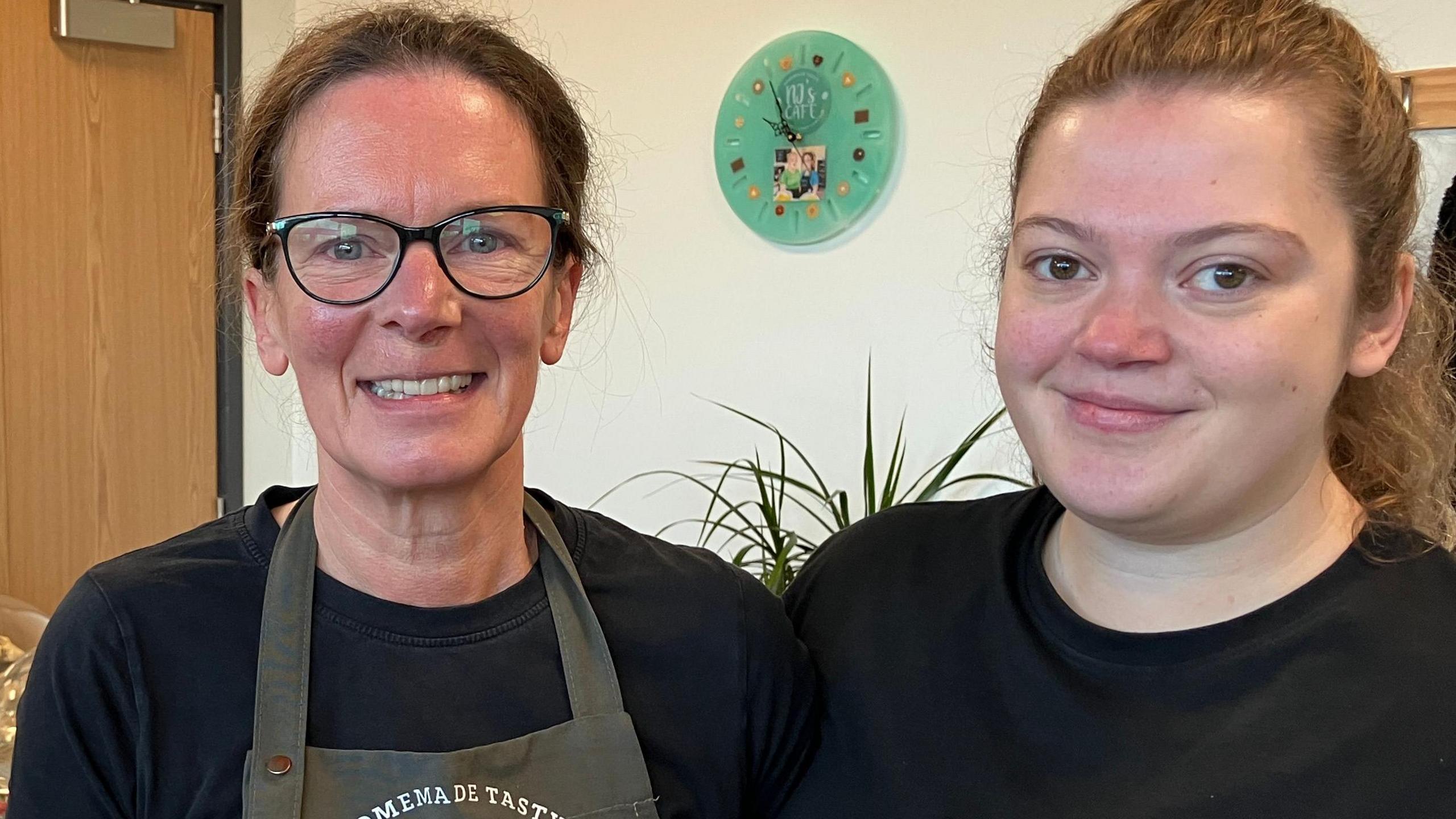 Jo Beattie with brown hair tied back, wearing black-rimmed glasses, a black top and a green overall, stands next to her daughter Nadia, who has light brown hair tied back and is also wearing a black t-shirt. 