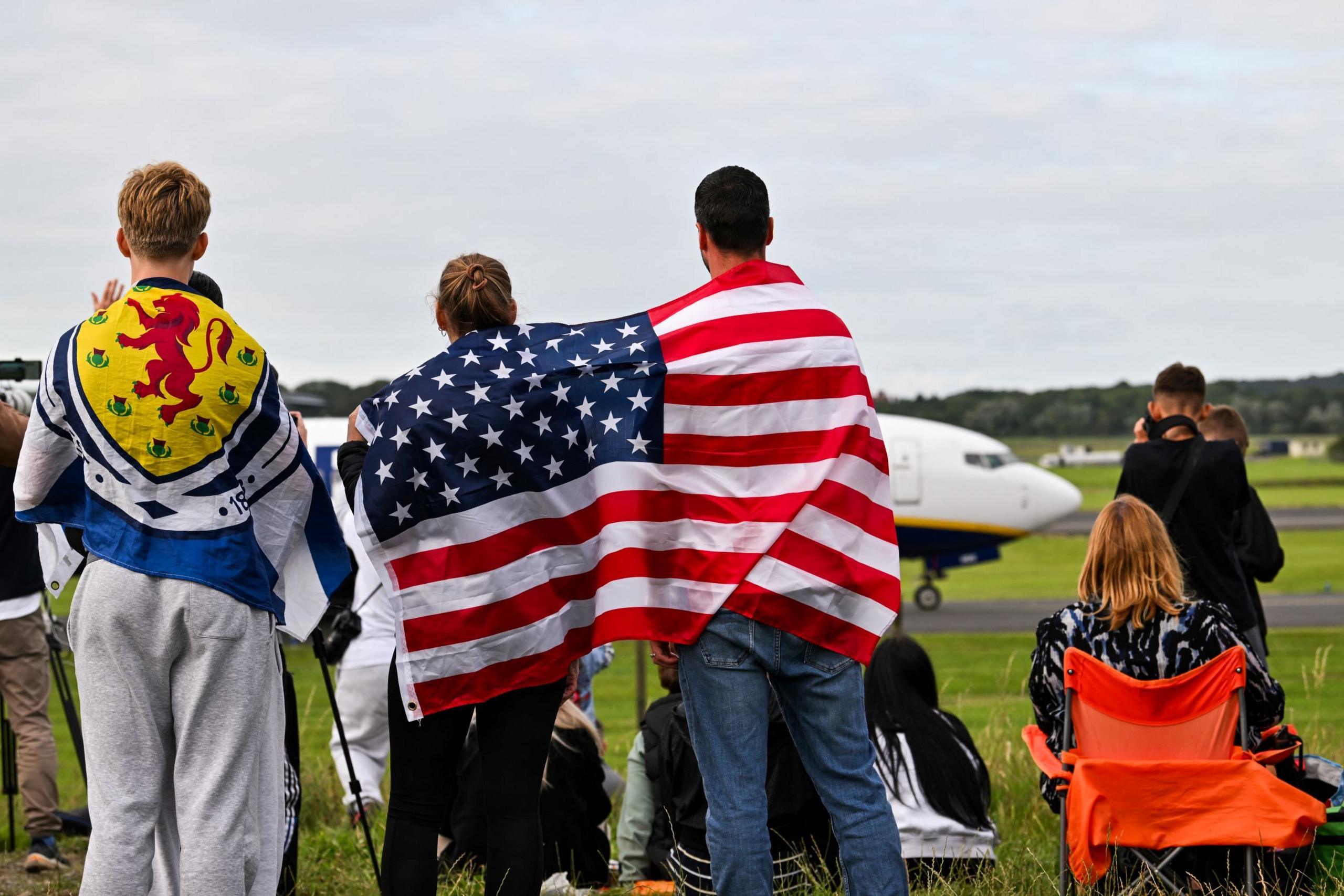 Trump supporters at airport watch his plane land. One has a Scotland flag on his back and two otghers have an American flag draped round their shoulders. Other people are sitting and standing looking at the runway.