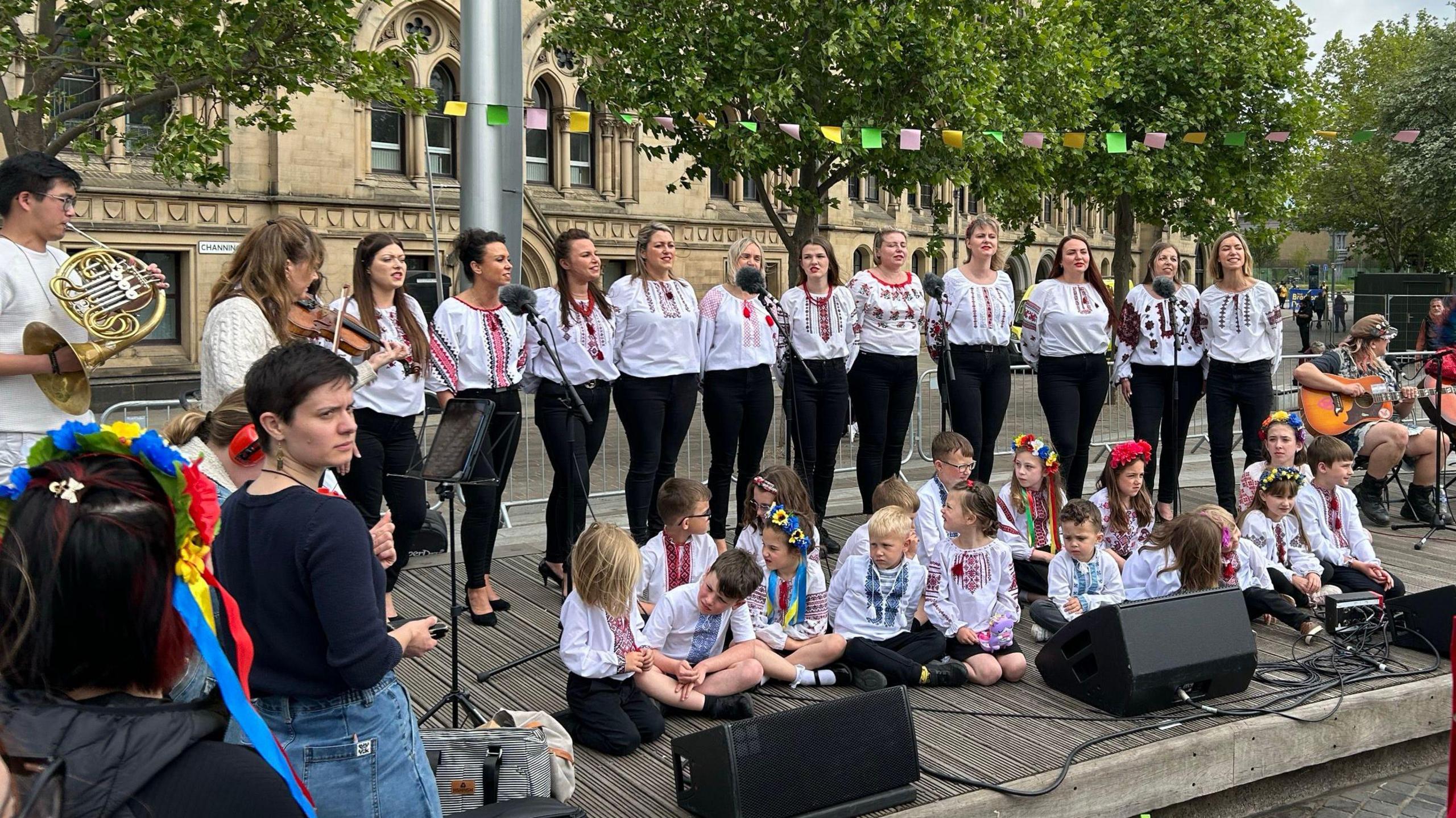 A line of women all wearing white singing outside the City Hall in Bradford. Children are sat in front of them and there are people playing guitars to the right of the image, and a person playing a violin and person playing a brass instrument to the left of the image.
