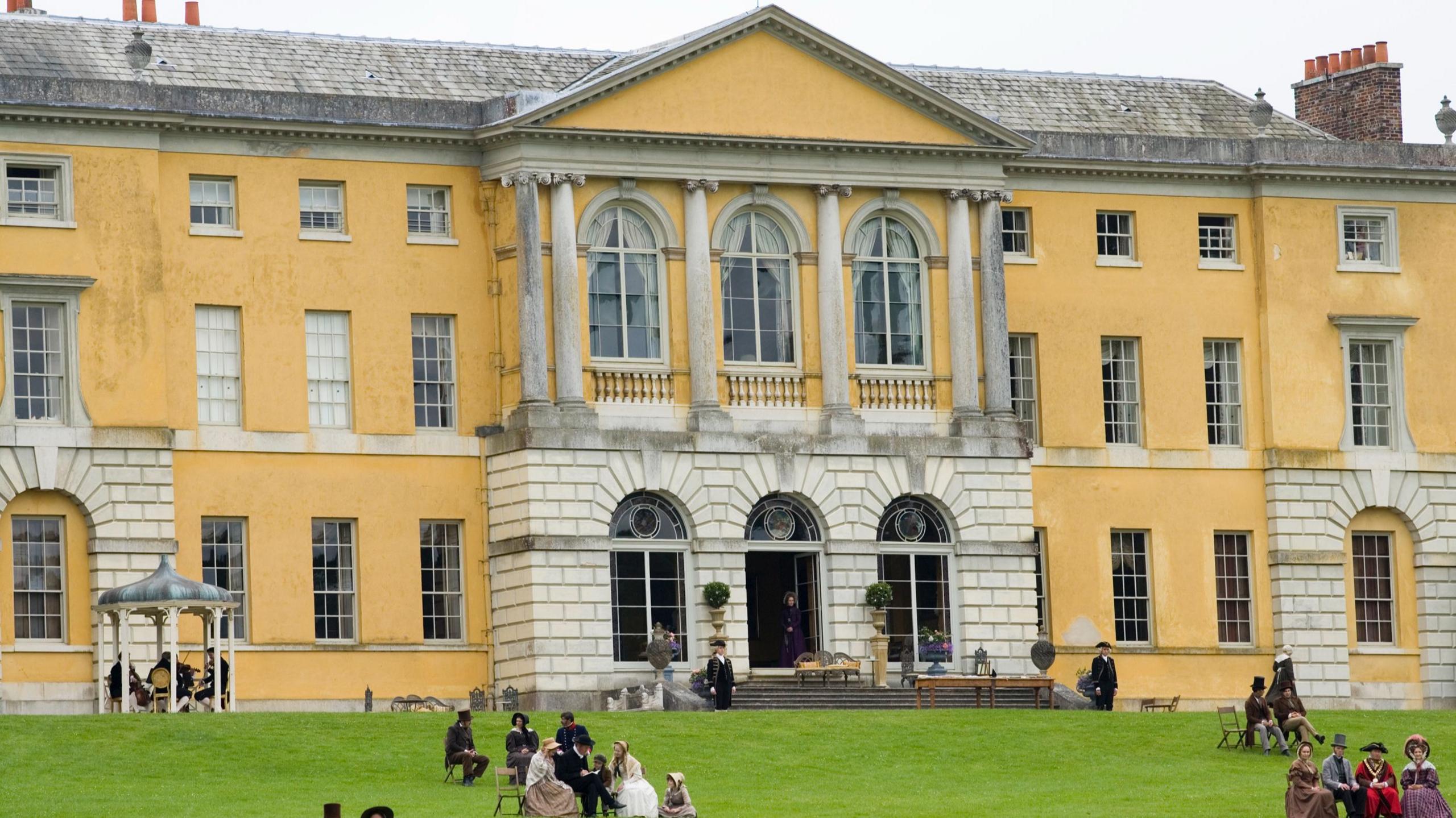 A grand yellow mansion with a grey roof stands prominently in the background, featuring arched doorways and tall columned windows. In the foreground, a group of individuals dressed in historical costume gather on the lawn.