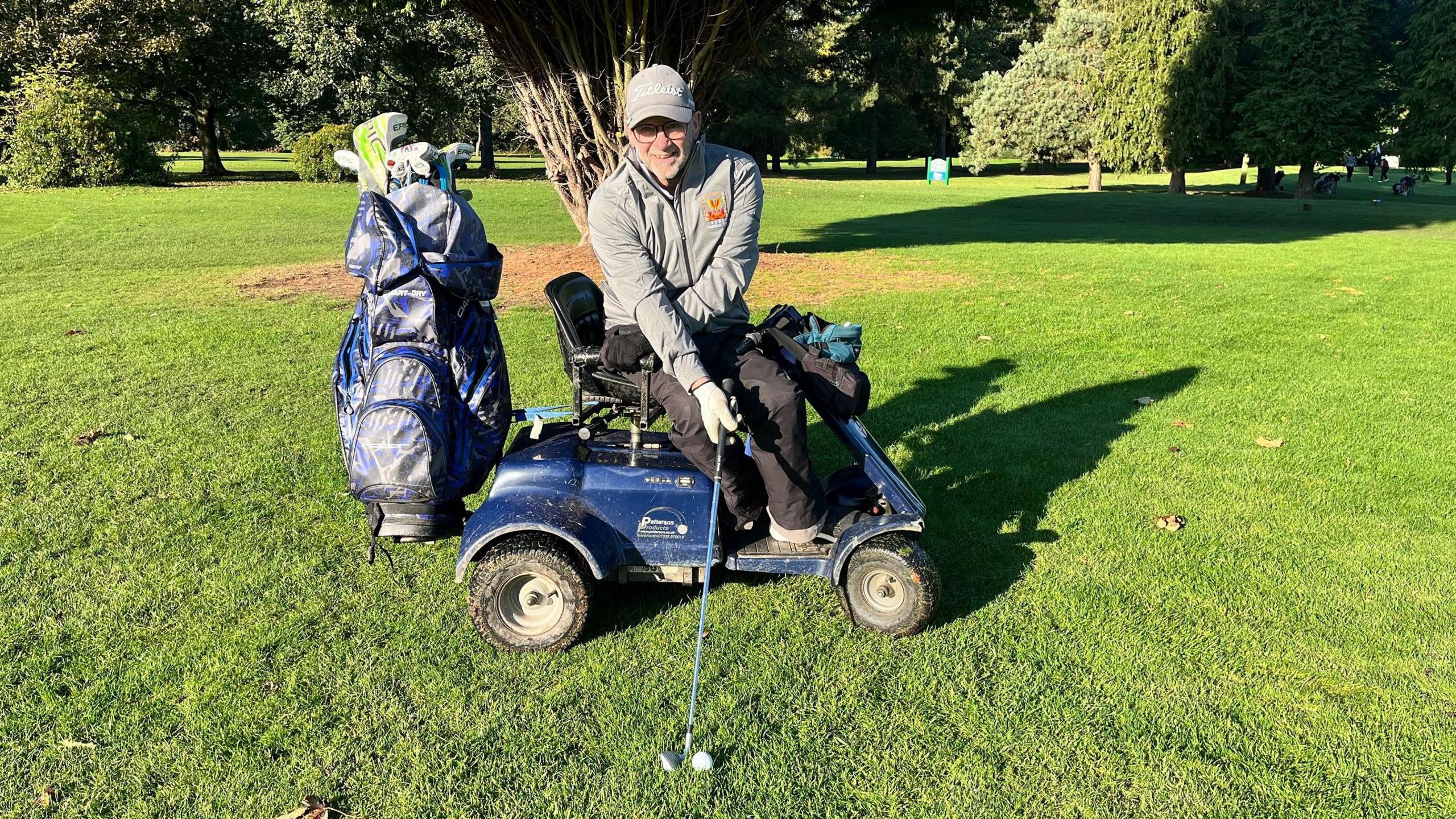 Terry Kirby prepares to strike a golf ball from the fairway while holding the club in one hand and sitting in a buggy. He is wearing a light-grey jacket,  matching cap and dark-grey trousers. The blue buggy carries a blue golf bag.