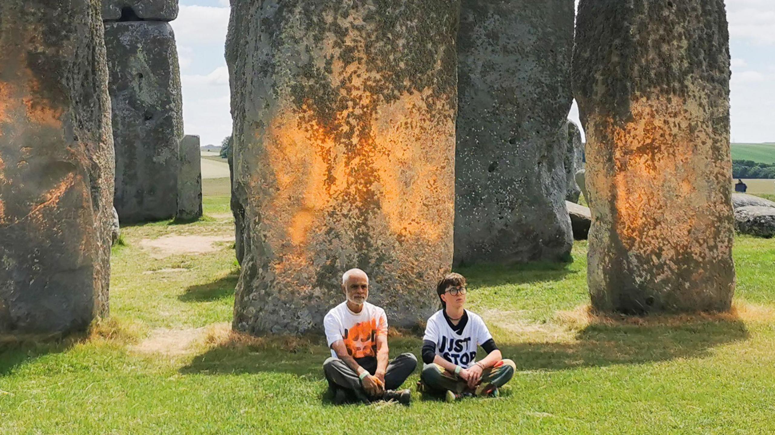 The Stonehenge monument on a bright sunny day. Some of the stones appear to have orange paint on them. Two of the protestors, Ms Lynch and Mr Naidu are sat in protest in front of the stones.