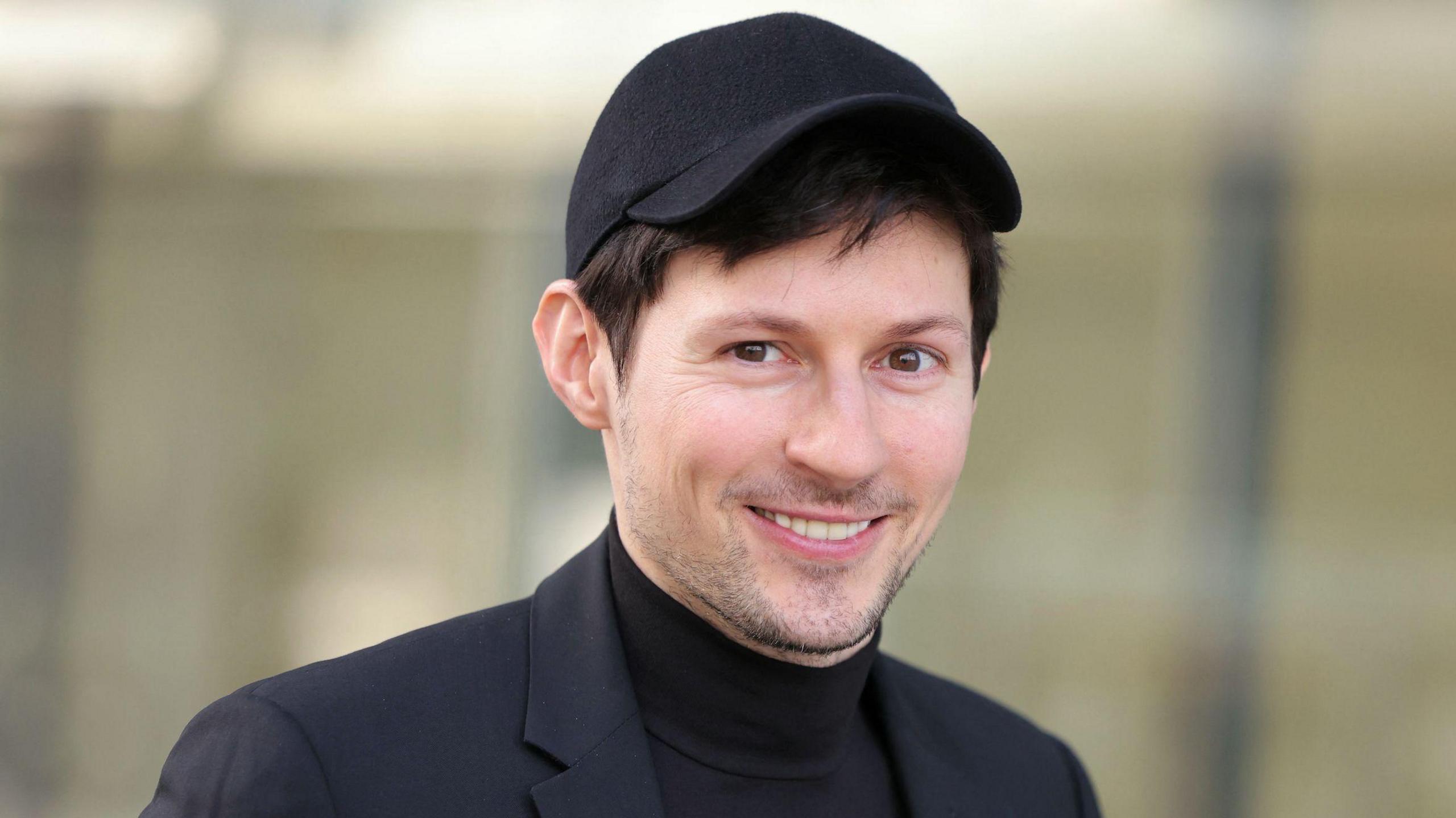 Pavel Durov, who has brown hair, brown eyes and light stubble and is wearing a black suit, a black T-shirt and a black baseball cap, smiling at the camera outside a Paris court for a hearing