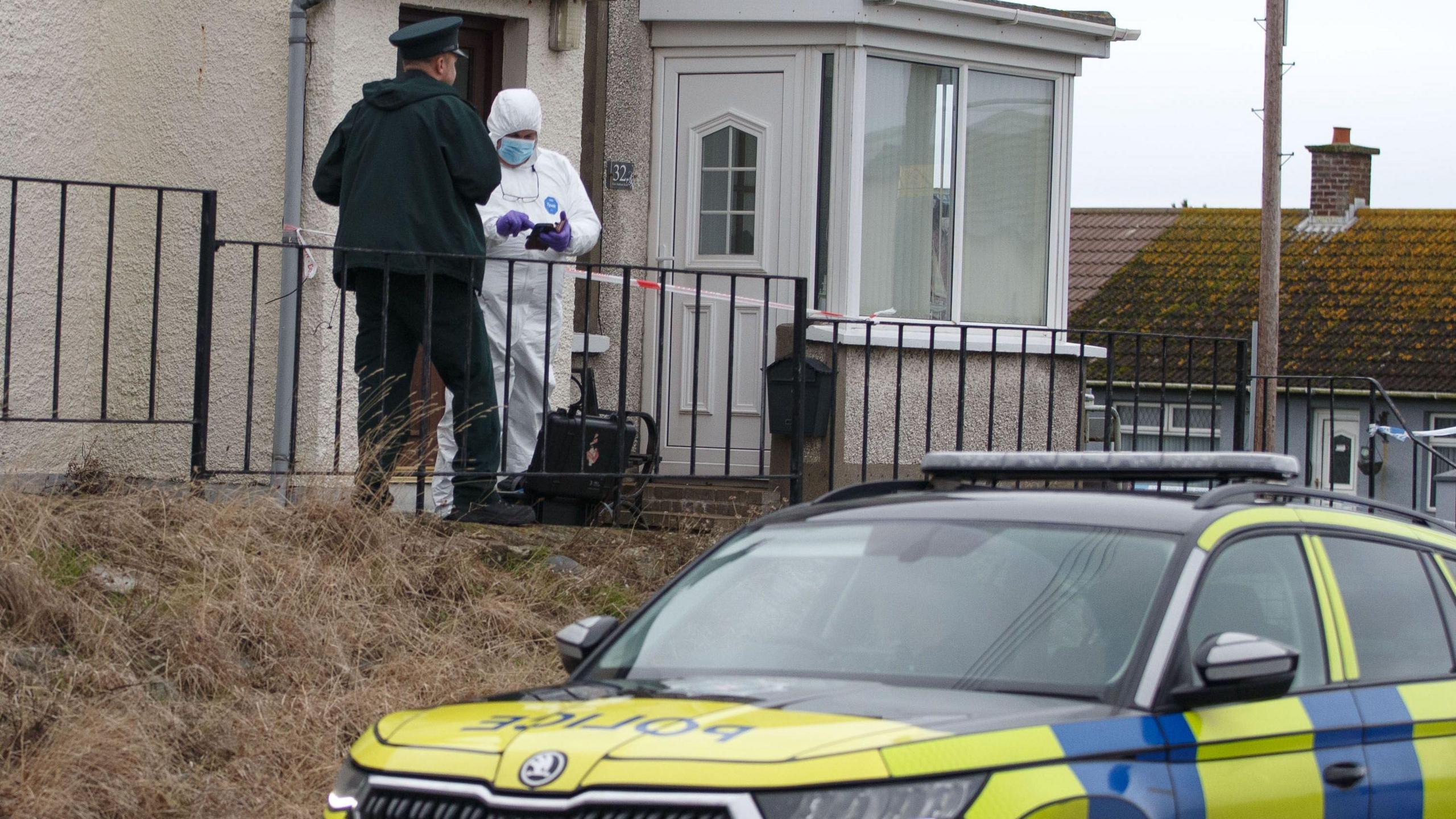 A police man and person in white PPE gear are standing outside a white pebble dashed house. A police car is parked in the foreground