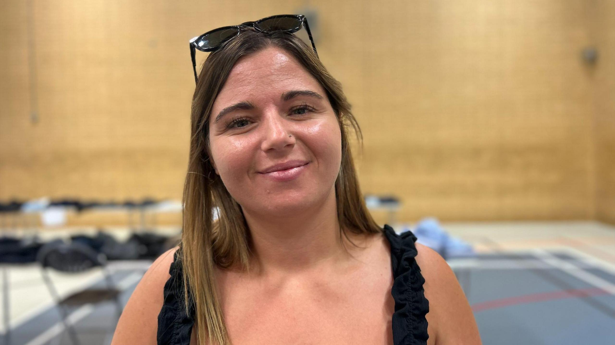 Natalie has long brown hair and has a pair of sunglasses balanced on her head. She has a black strappy top on and smiles for the camera.