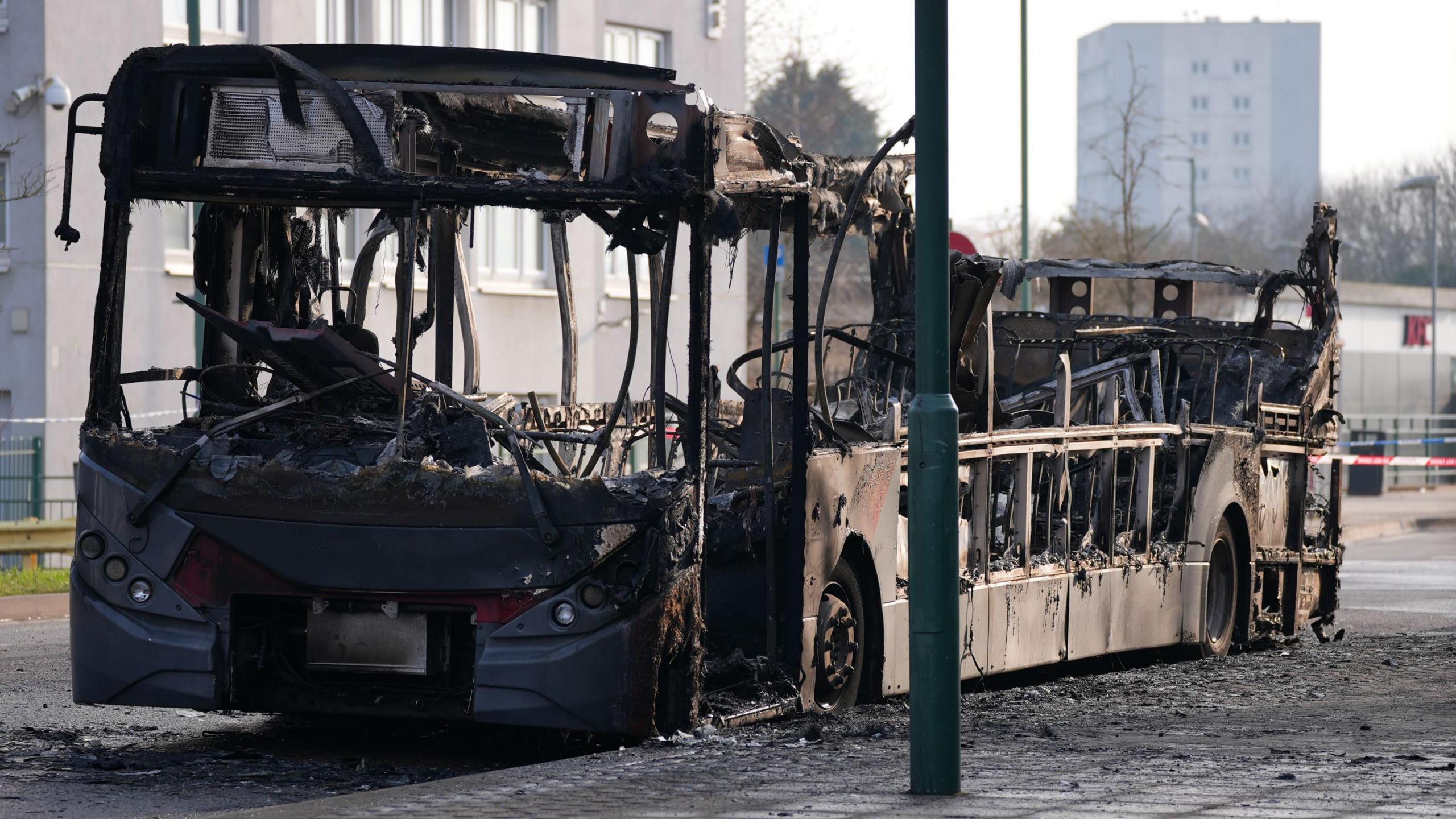 A burnt-out bus without a roof, parked on the side of a road. Mangled, charred metal sticks out from what remains, with no windows or windscreen