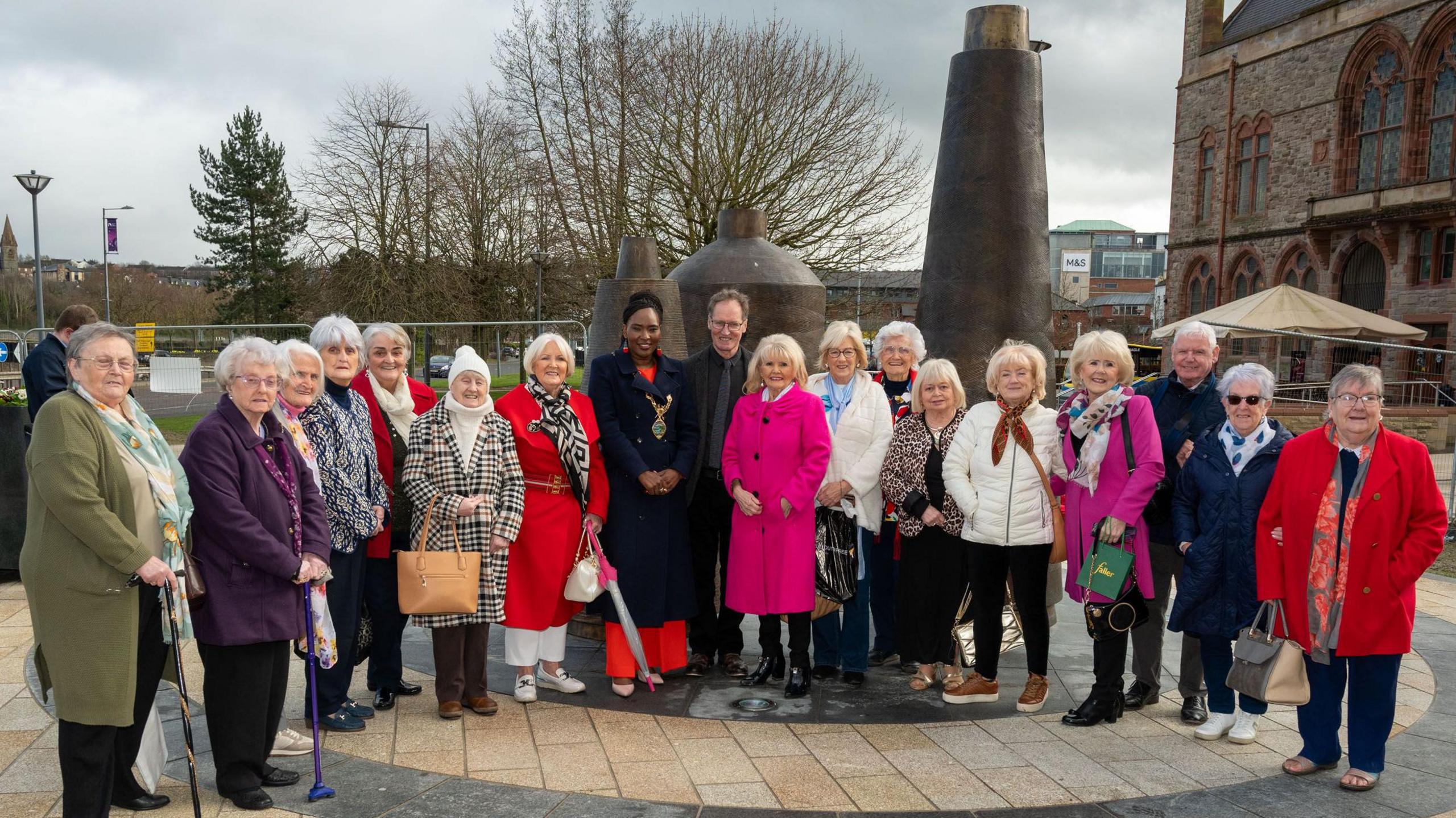 A group of people stand in front of a piece of public art. The majority are women, dressed in winter coats. A town hall is behind them.