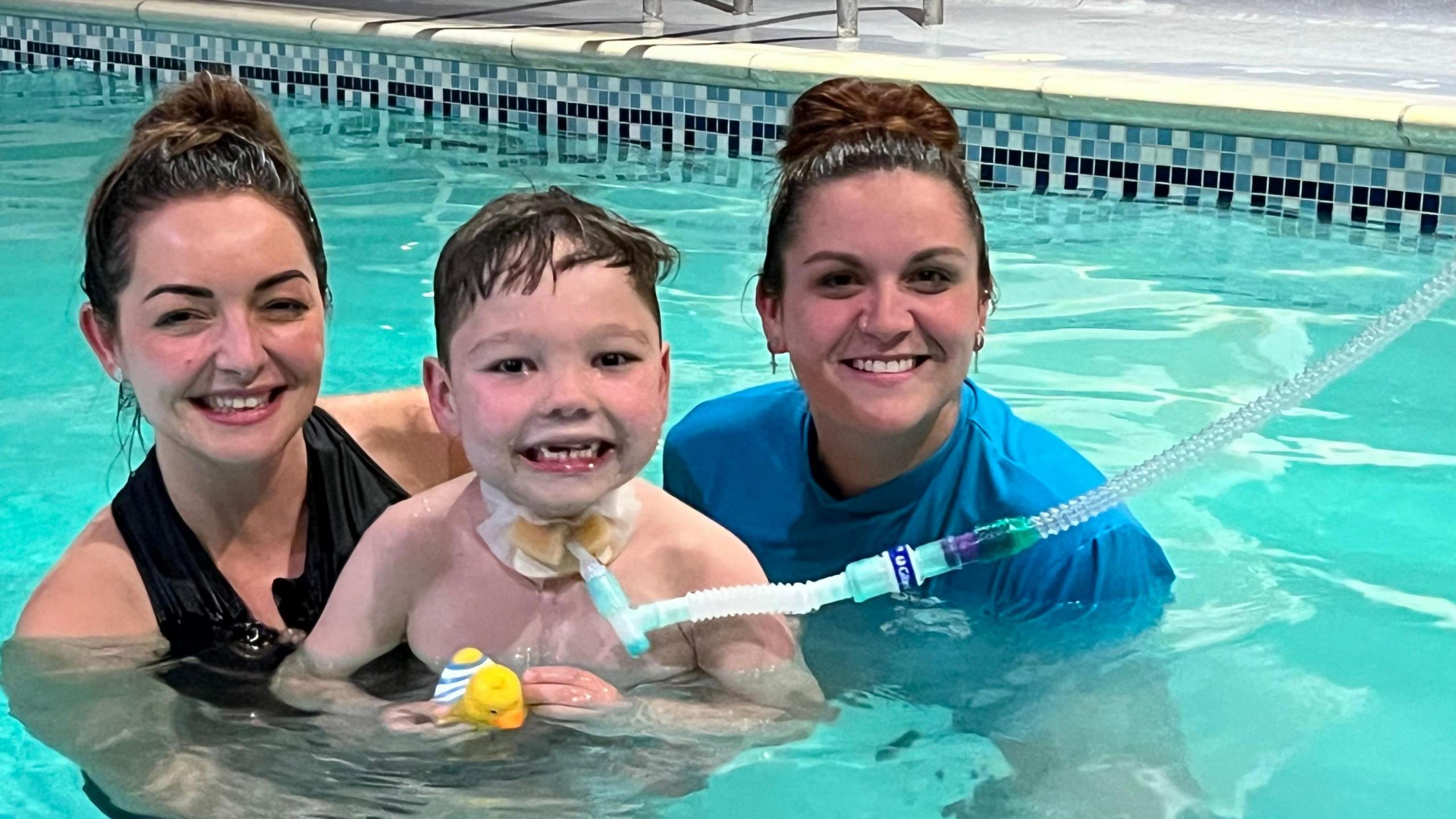 Mum Shevonne and teacher Kayleigh support Henry, six, in the blue water of a swimming pool. All three are smiling. Henry has a tube attached to his neck which runs up towards the side of the pool. Shevonne wears a black swimsuit, while Kayleigh wears a blue T-shirt.