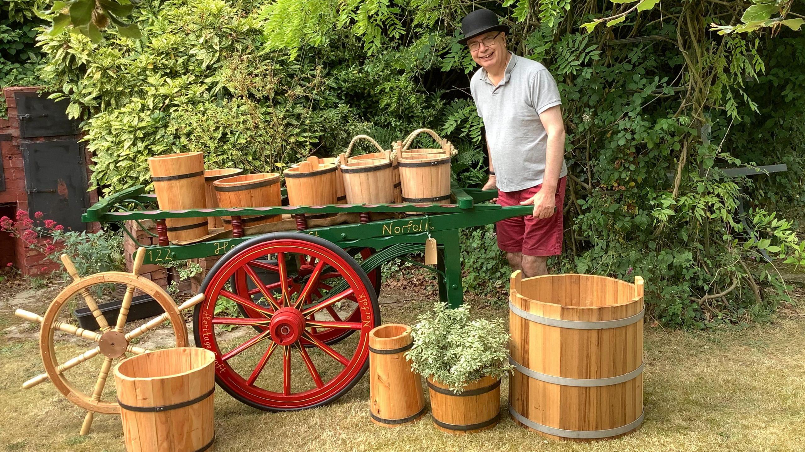 Alan Paulus wearing shorts, a T-shirt and a black bowler hat holding a large green cart, which has red wheels. On top and around the cart are buckets of all sizes.