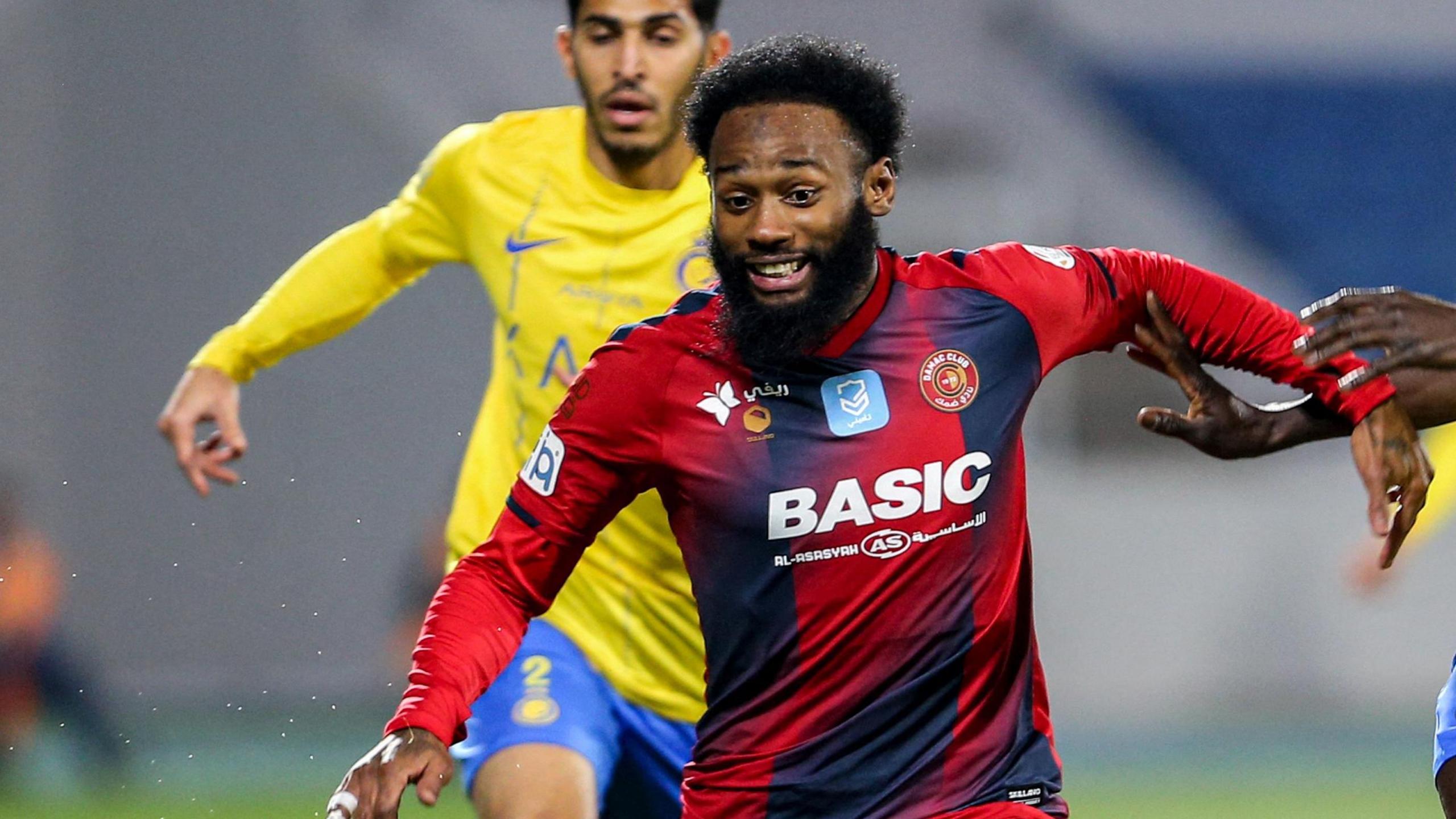 Footballer Georges-Kevin Nkoudou, wearing a red and dark blue Damac shirt, strains in concentration as he tries to shrug off the attentions of a defender who is out of shot, with another defender in yellow and blue chasing in the background