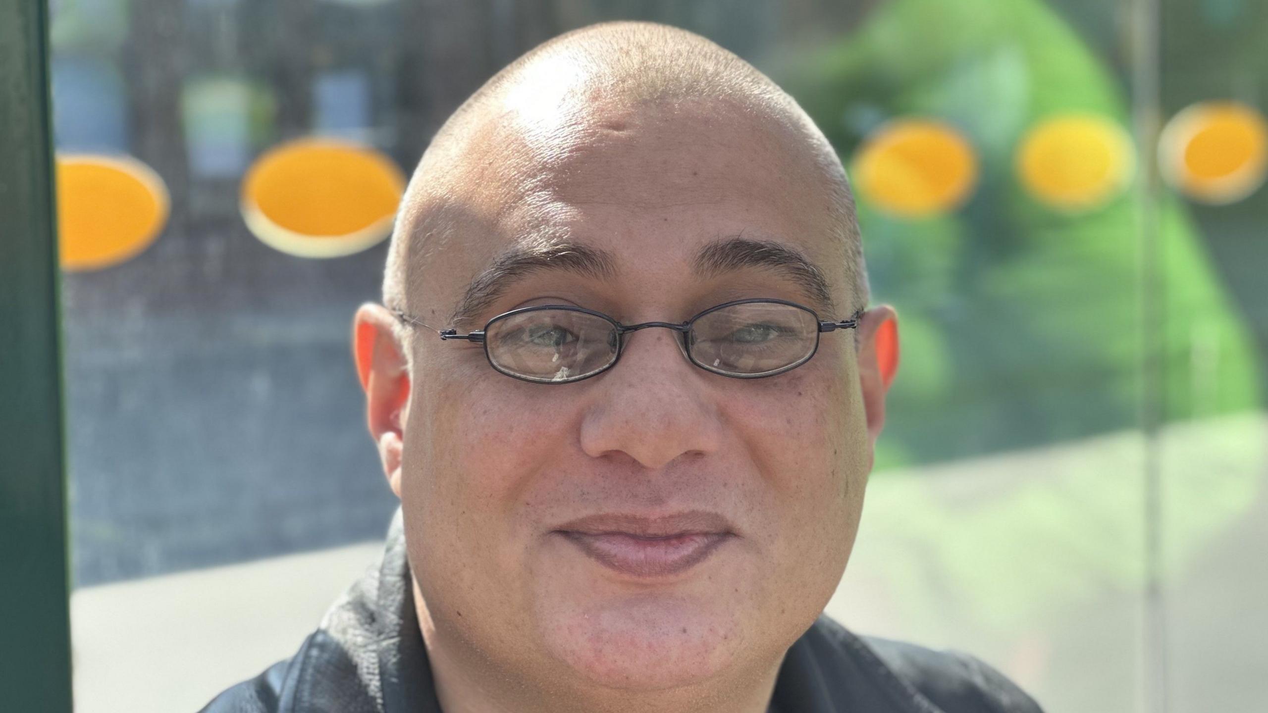 Darrell Dragon smiles at the camera while sitting in a bus shelter. He is bald and wears glasses. He wears a black leather jacket. 