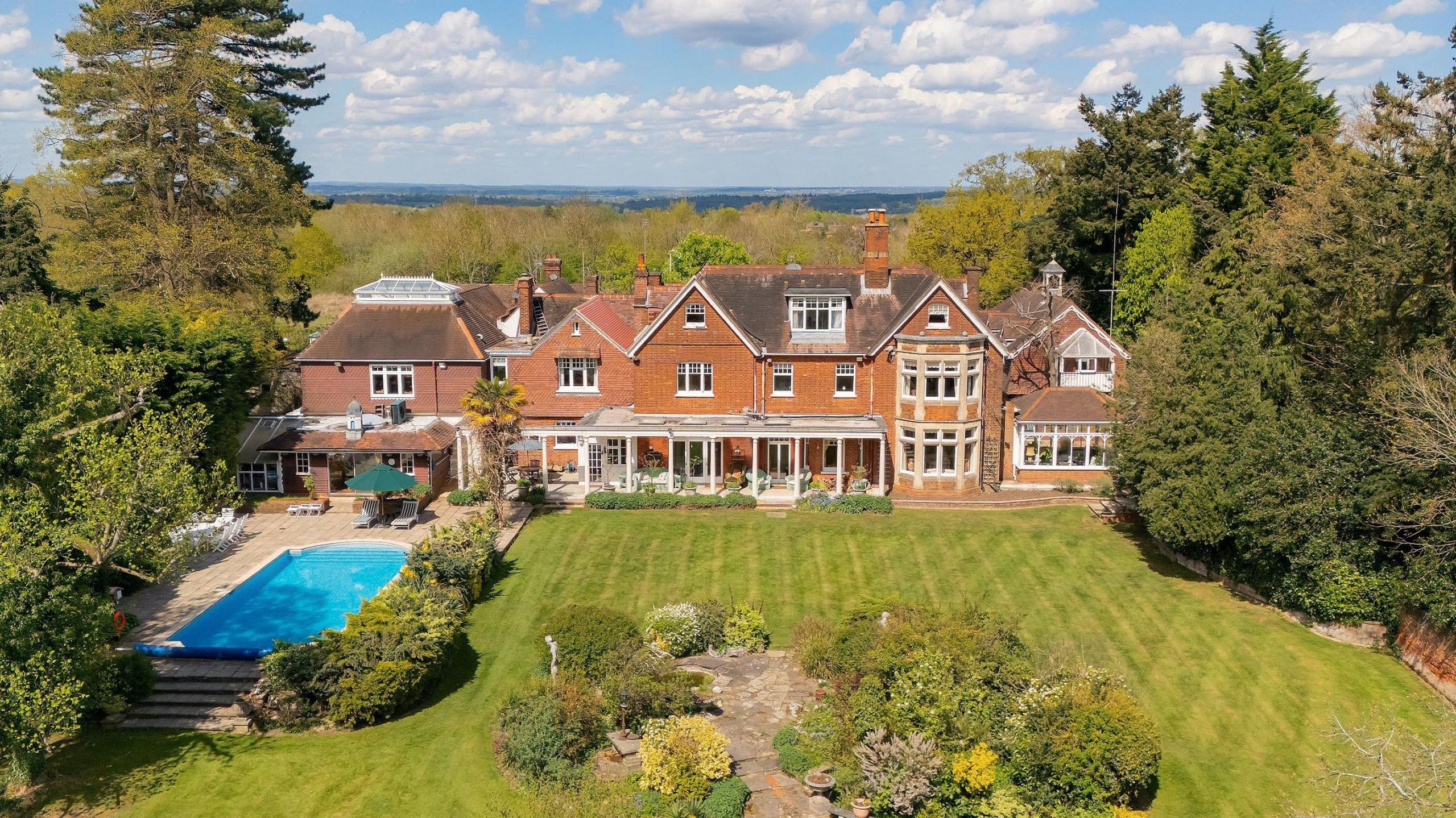 A huge house can be see from a drone. It has a well kept green lawn and is surrounded by huge trees. There is also a swimming pool on the left hand side. The middle of the huge house has columns to hold up a roof creating an outdoor seating area under cover.