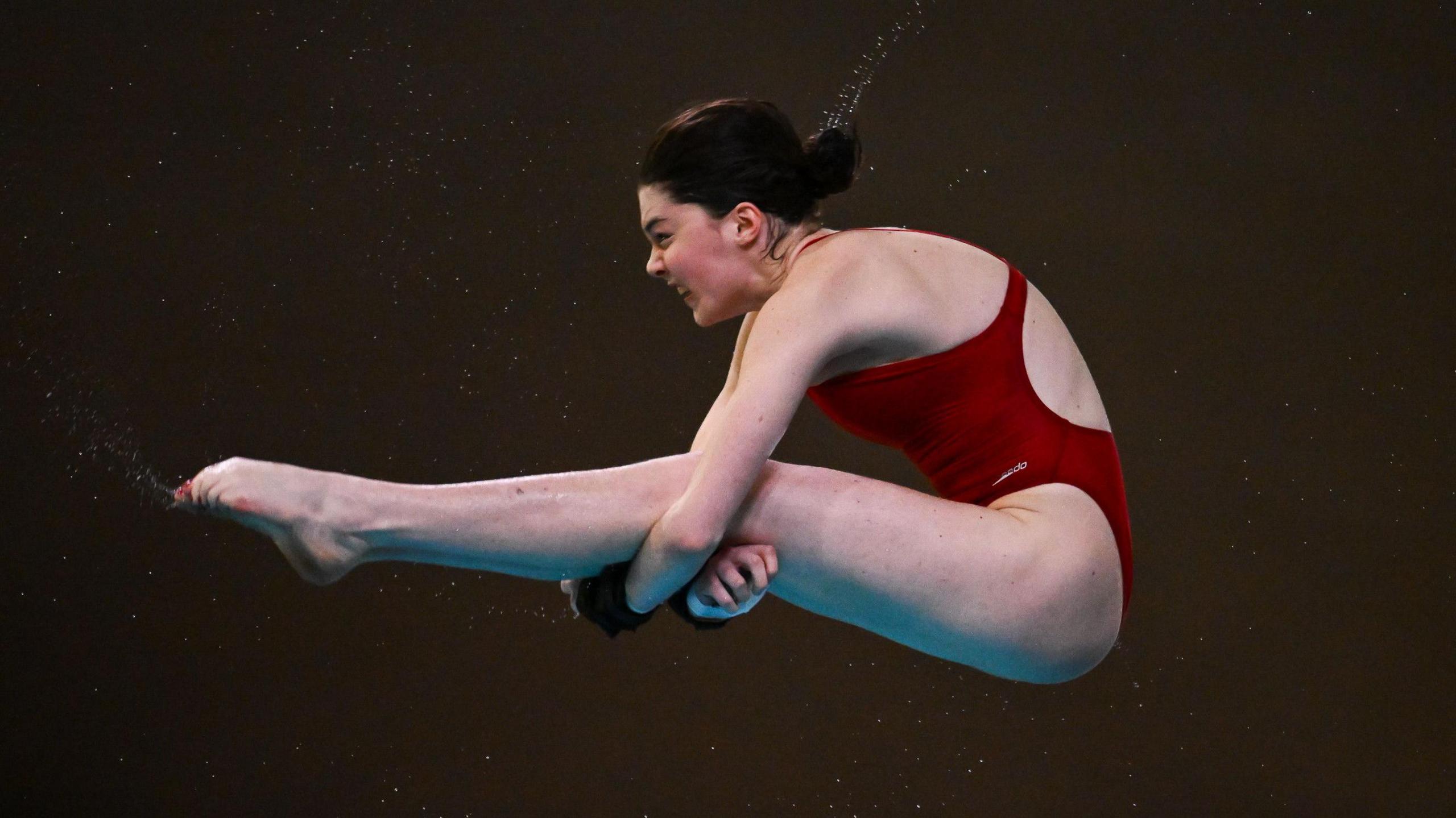 Andrea Spendolini-Sirieix is mid-dive with her legs out stretched with her arms tucked beneath her legs. She is wearing a red swimsuit.