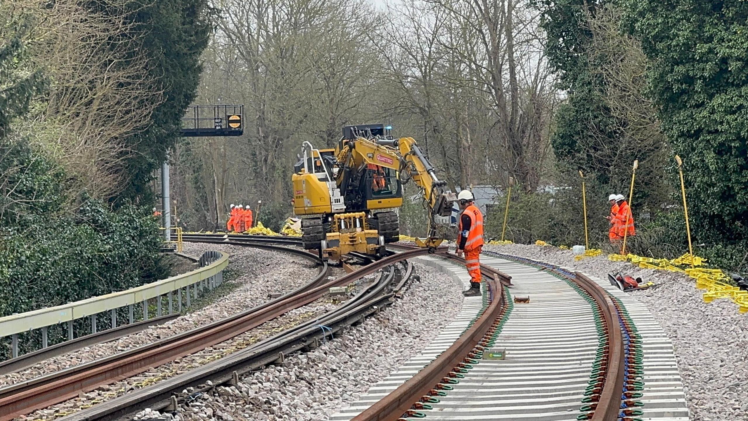 People in orange hi-vis outfits stand on a railway line as a machine lifts pieces of track away from the floor