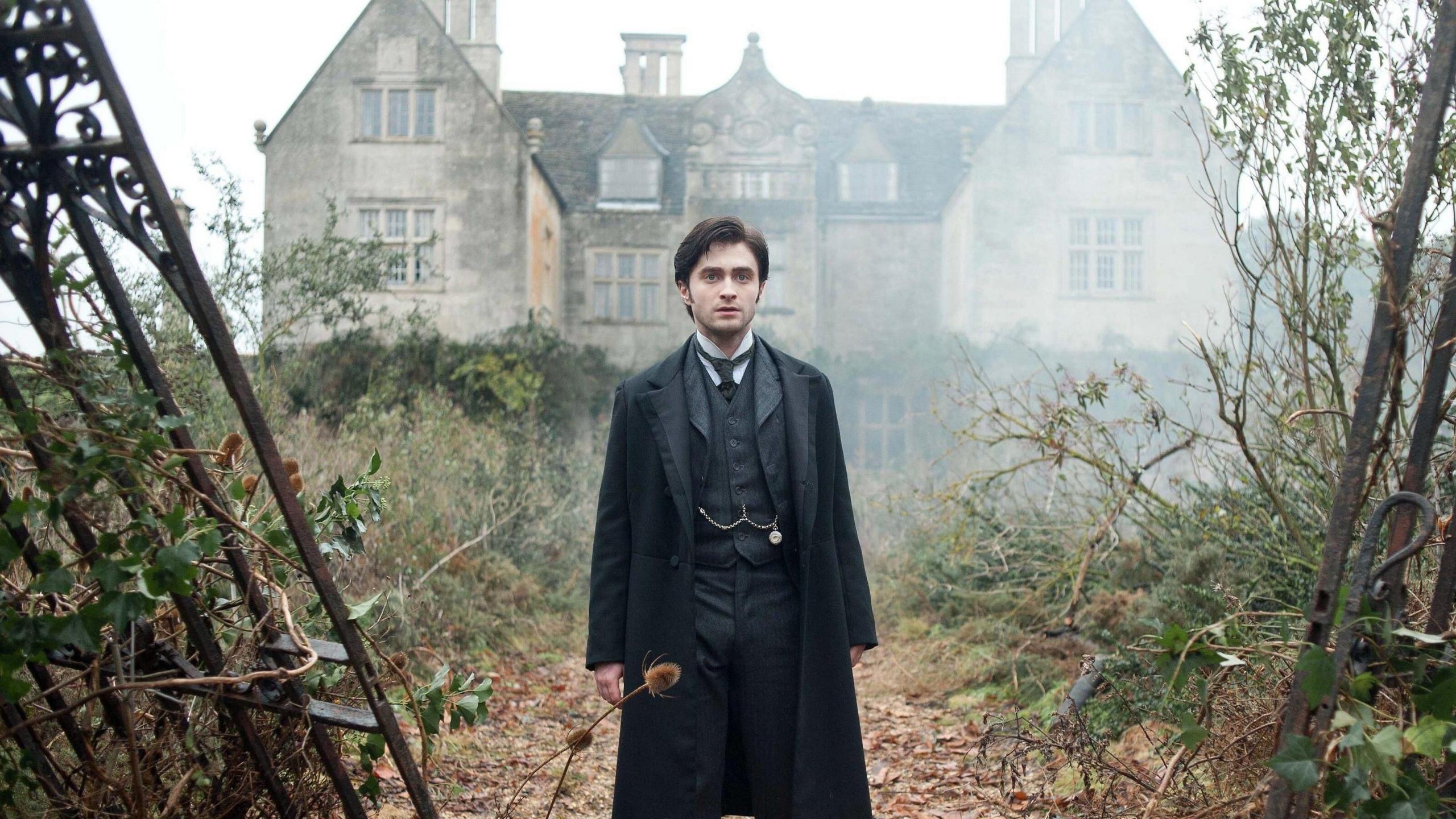 Daniel Radcliffe stares into the distance, dressed in a black suit, waistcoat with a chain across the middle and overcoat. He is in an foggy, overgrown garden in front of an old grey stone building. To his sides are the edges of what looks like wrought iron gates that have fallen off their hinges.