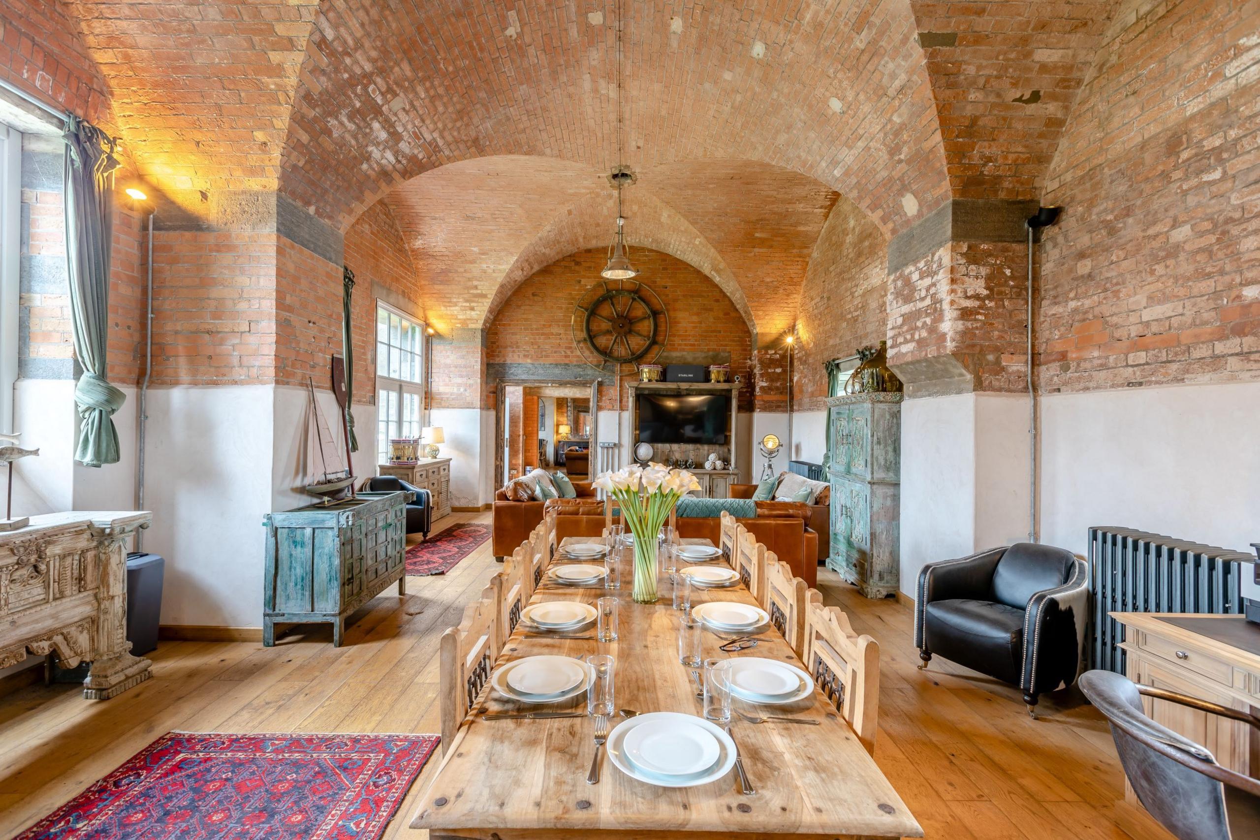 A view of the living room from the other end of the dining table. The impressive high ceiling is domed, with a large ship's wheel mounted on the wall. Red Afghan rugs cover parts of the wooden floor, with a leather armchair in one corner. 