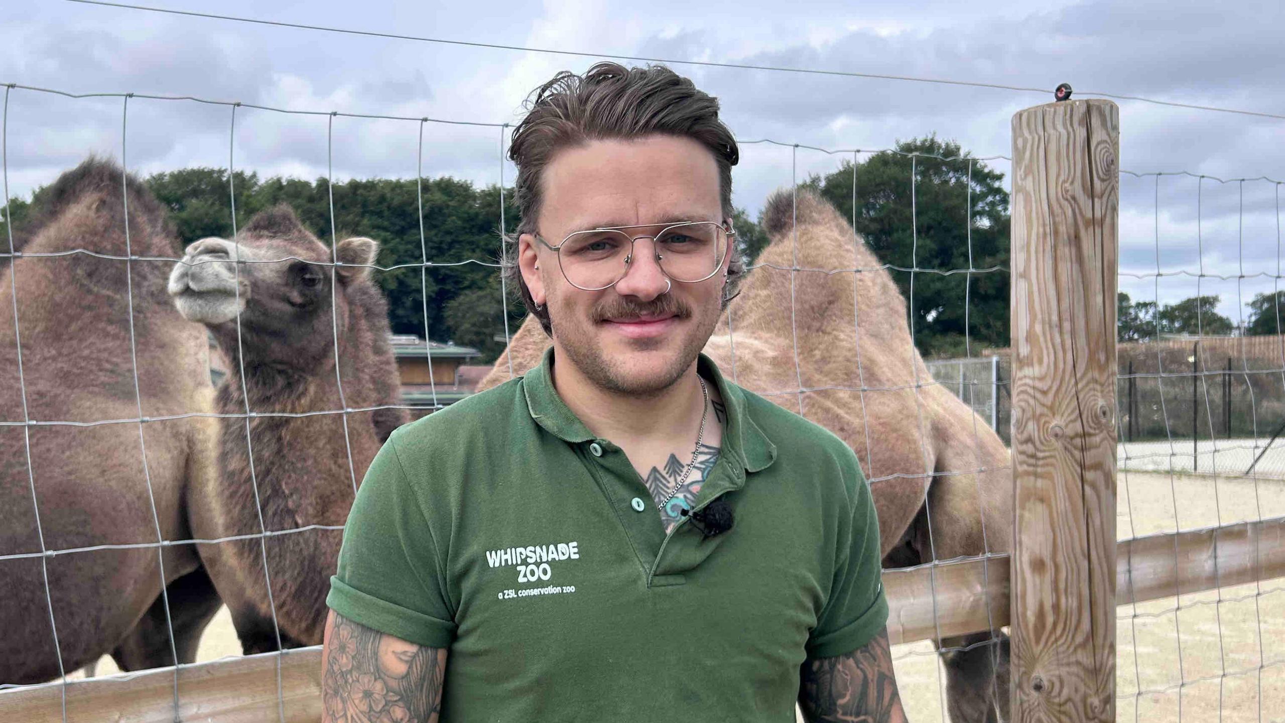 George Spooner with short brown hair and beard wearing a green Whipsnade Zoo T-shirt and standing in front of a wire and wood fence, behind which are two camels. There are trees in the background.