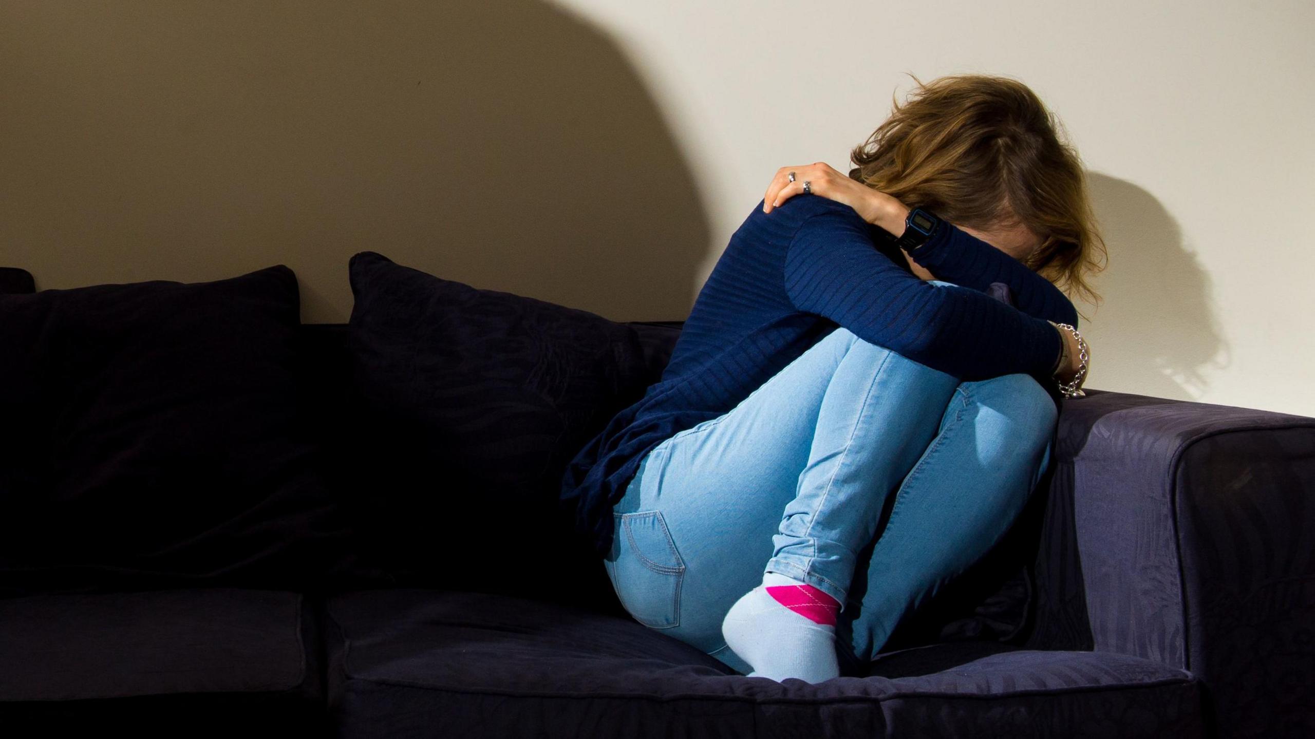 A woman covers her face with her arms as she cowers on the corner of a sofa