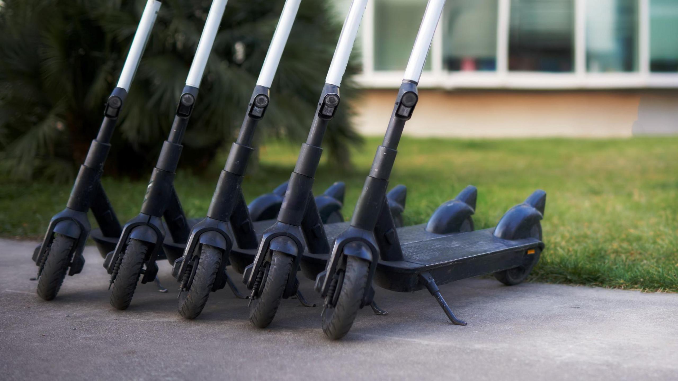 A row of five black scooters parked on a pavement next to a grassy area