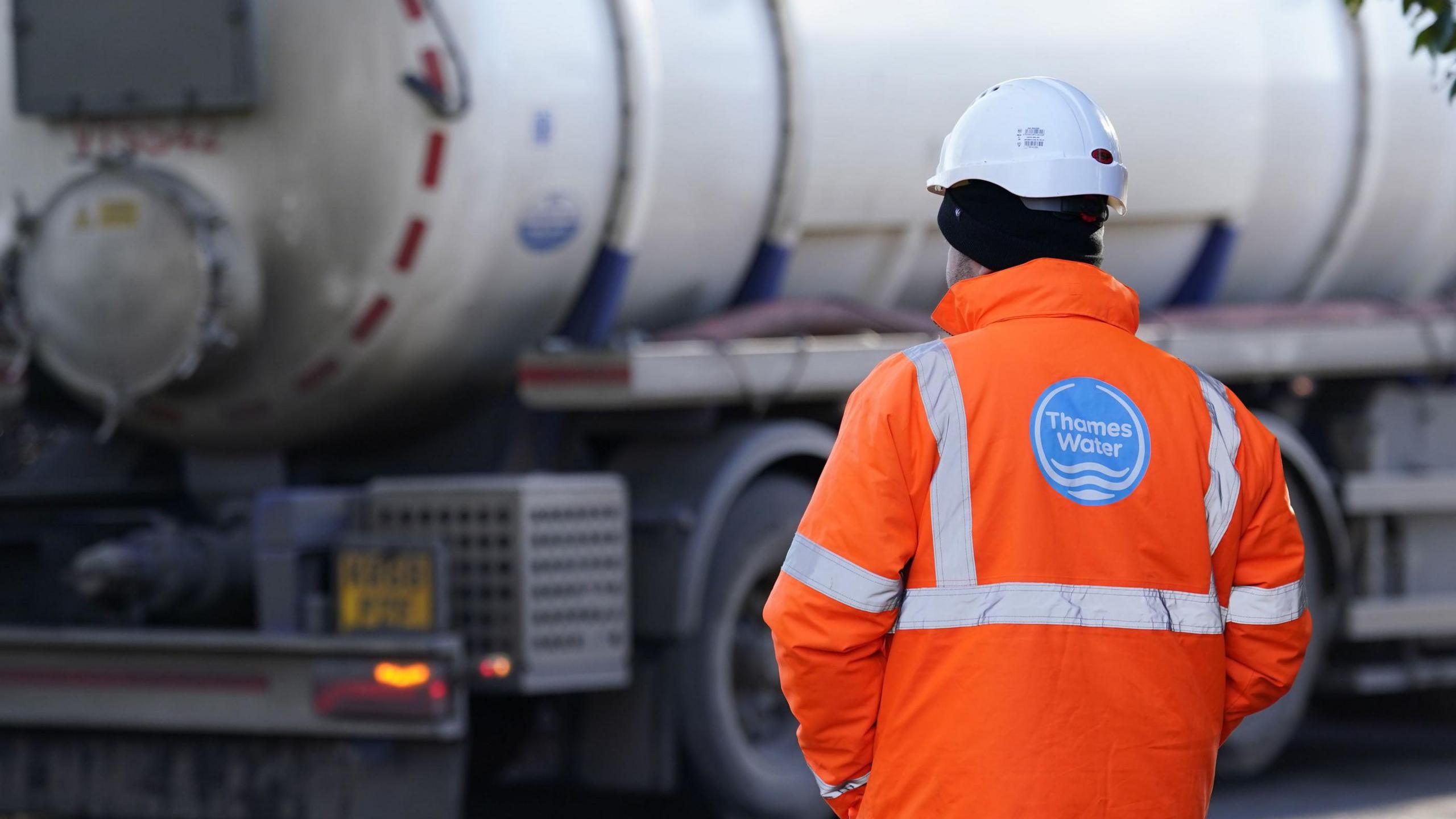 A Thames Water worker wearing a hard hat and orange jacket looks on as a lorry drives by