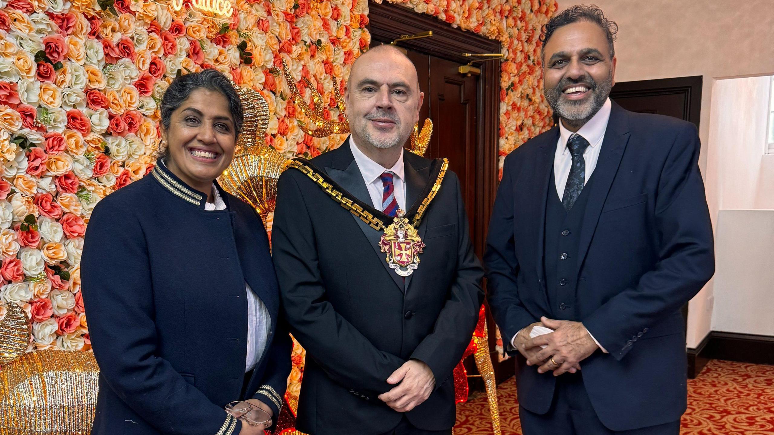 A woman and man stood either side of the mayor who is wearing a mayoral chain, suit and tie.  The woman, who is on the left, is smiling and wearing a blue jacket. The man, who is on the right of the mayor, is also smiling and is wearing a blue suit and tie. There is flowery wallpaper in the background. 