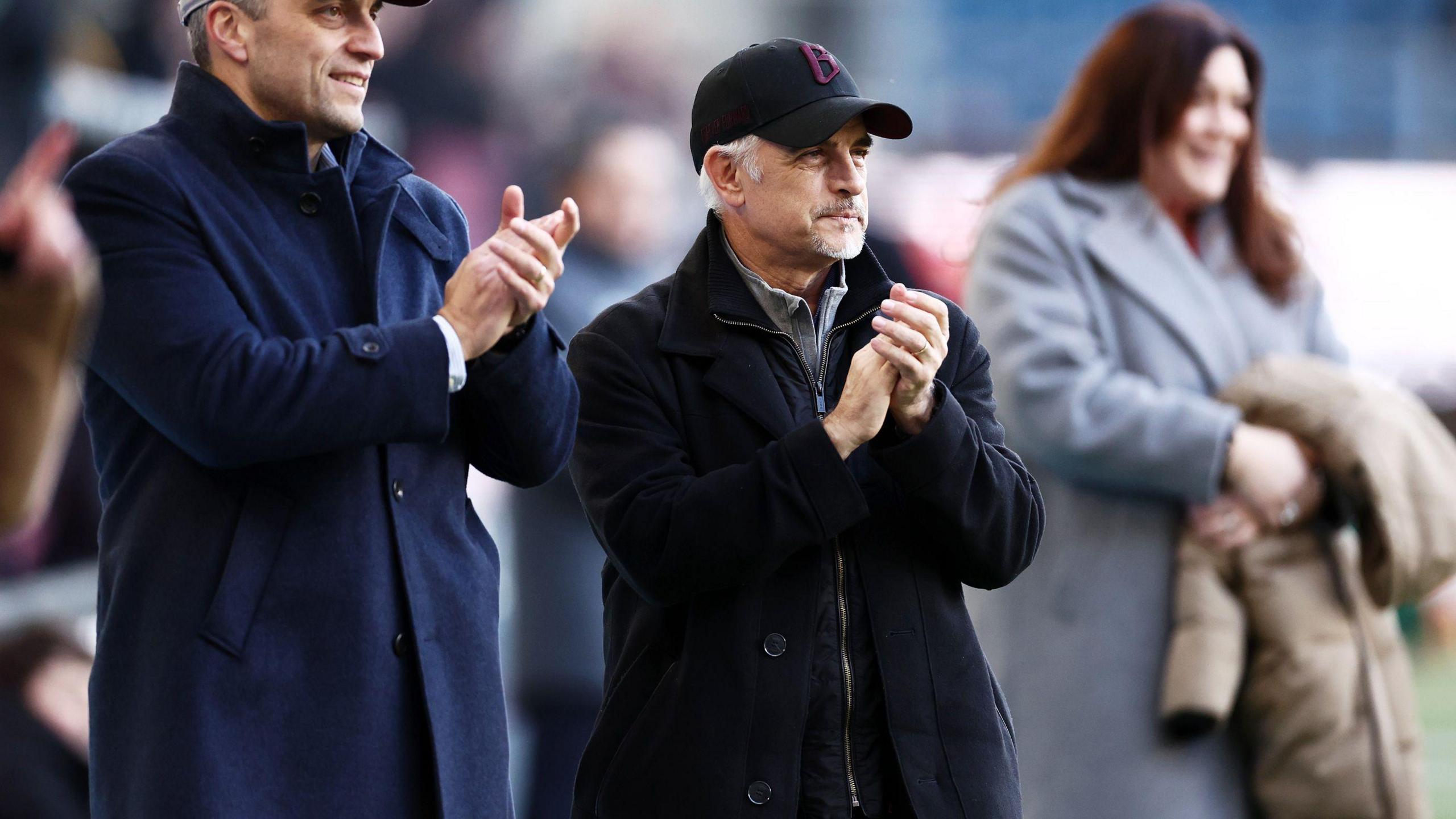 A photo of Burnley owner Alan Pace, wearing a black cap and black coat, applauding during a game at Turf Moor in February 2024