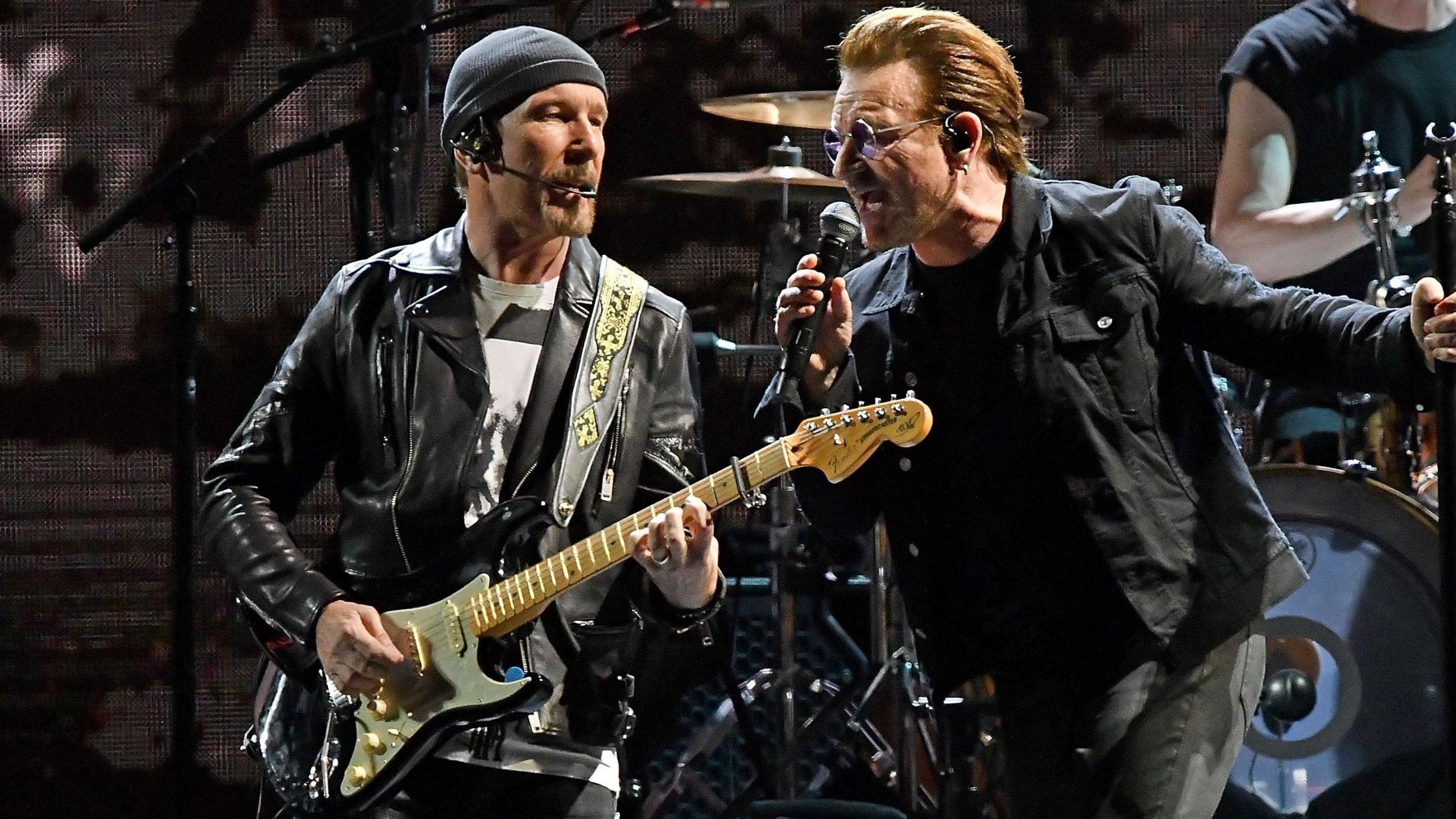 The Edge is onstage with Bono. He's wearing a black leather jacket and playing a guitar. Bono is singing into a mic.