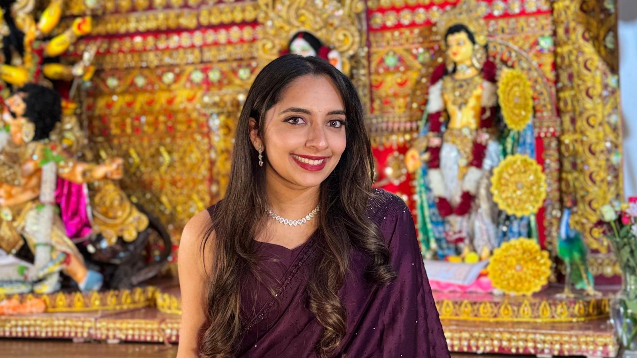 Vedika is dressed in a dark purple saree wearing her hair in loose curls and smiling. She stands in front of a Hindu shrine.