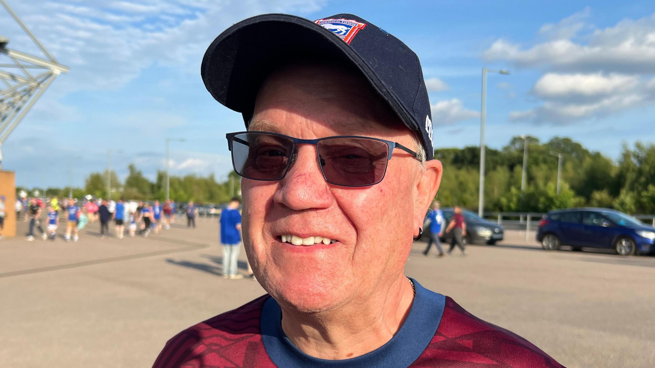 Peter Franklin stands in the middle of the picture looking towards the camera smiling. He is wearing a navy blue baseball cap, black sunglasses, and a maroon coloured Ipswich Town away shirt. Behind him are people walking past and rows of trees.