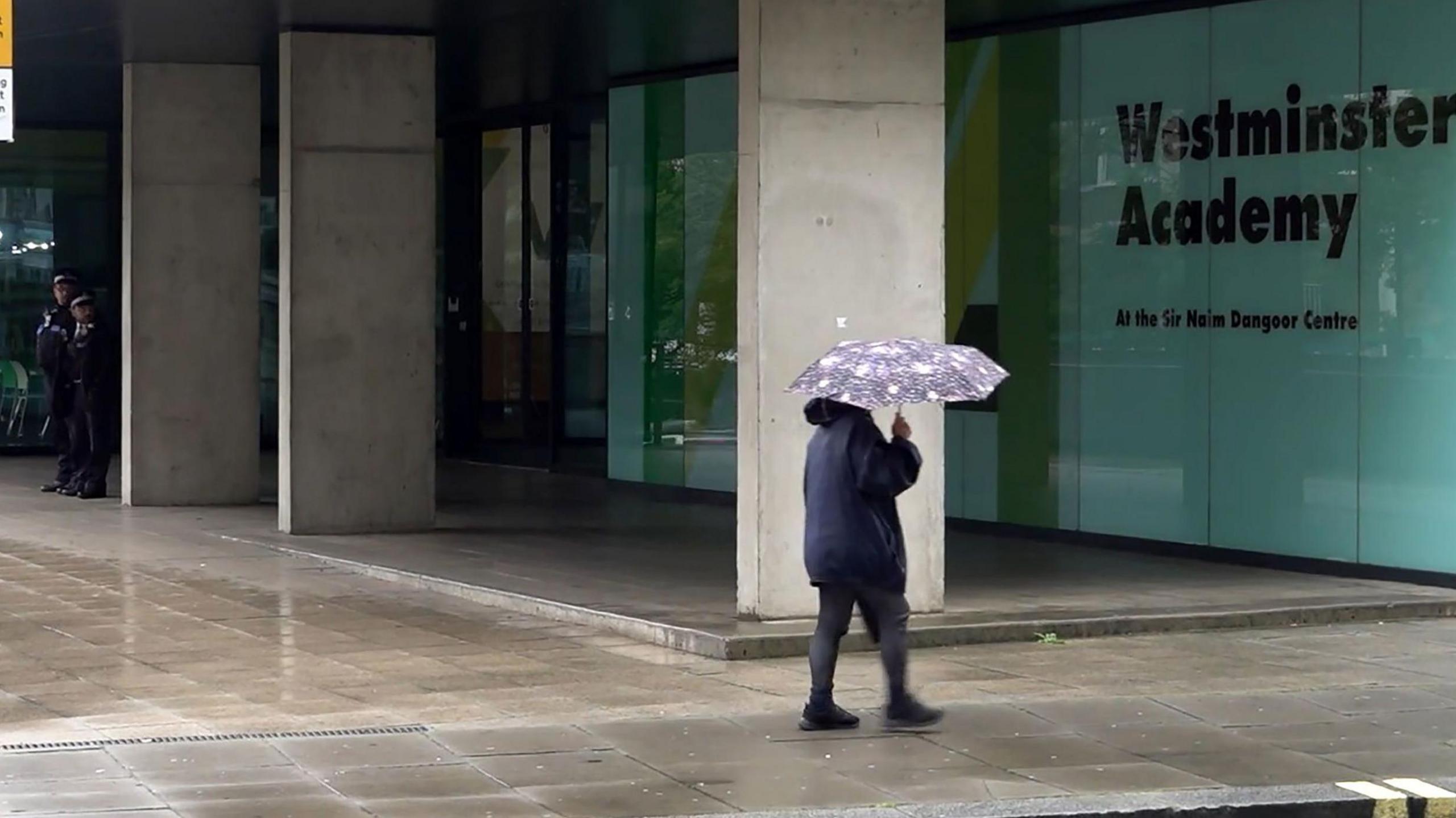 Two police officers stand guard outside Westminster Academy. A pedestrian holding an umbrella walks along the rain-soaked pavement