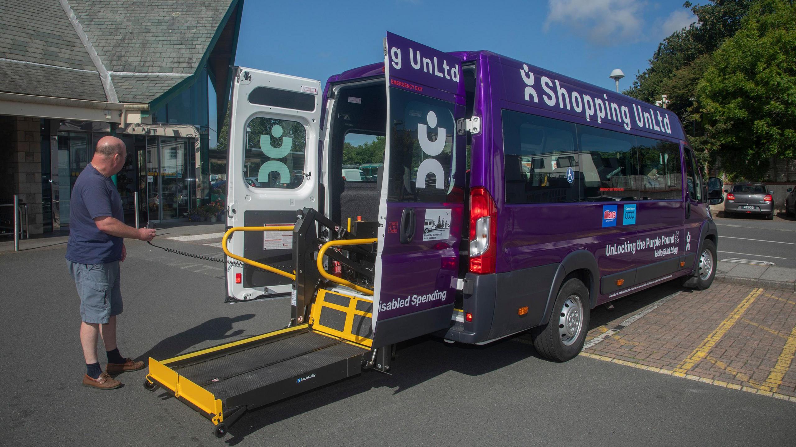 A purple Shopping UnLtd branded minibus is parked in a yellow hatched parking bay in a car park. Its back doors are open and there is a flat bed of a lift lowered to the ground. A man wearing a T-shirt and shorts is using a hand-held control box to operate the machinery.