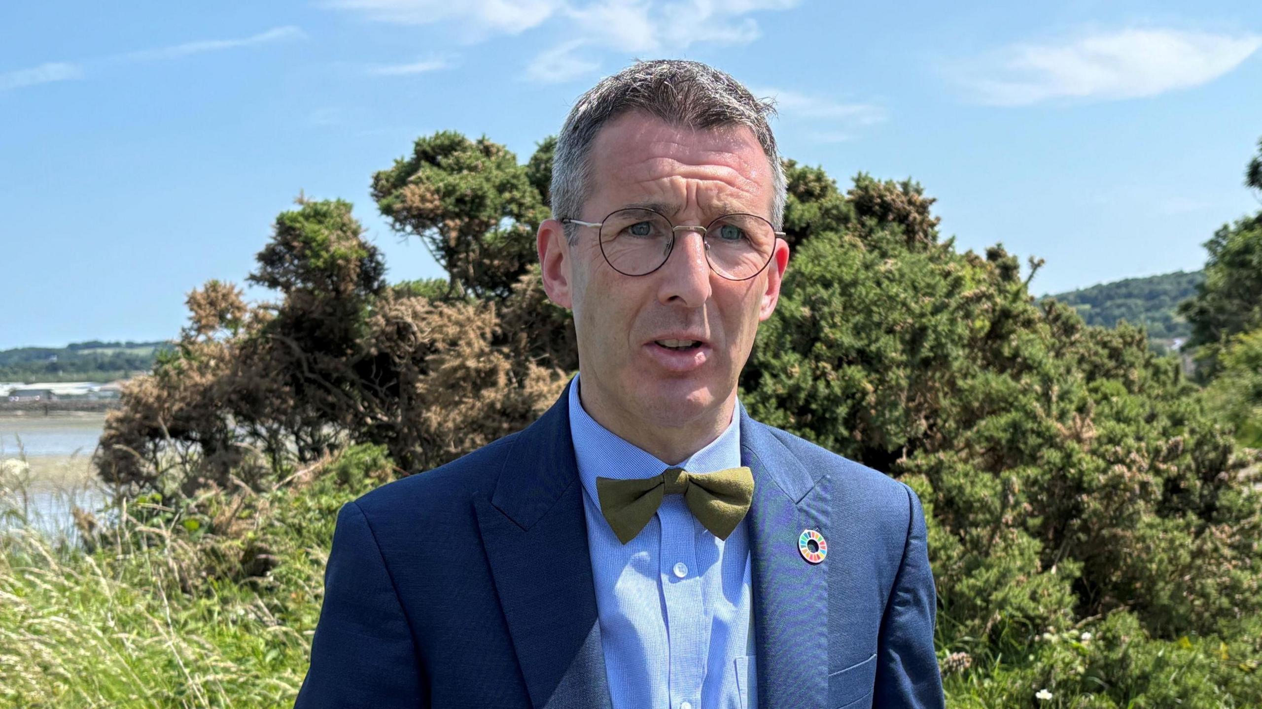 Andrew Muir wears a navy suit with a blue shirt and green bow tie. He has short grey hair and wears wire-rimmed glasses. He is standing on a path surrounded by grass and shrubs. 