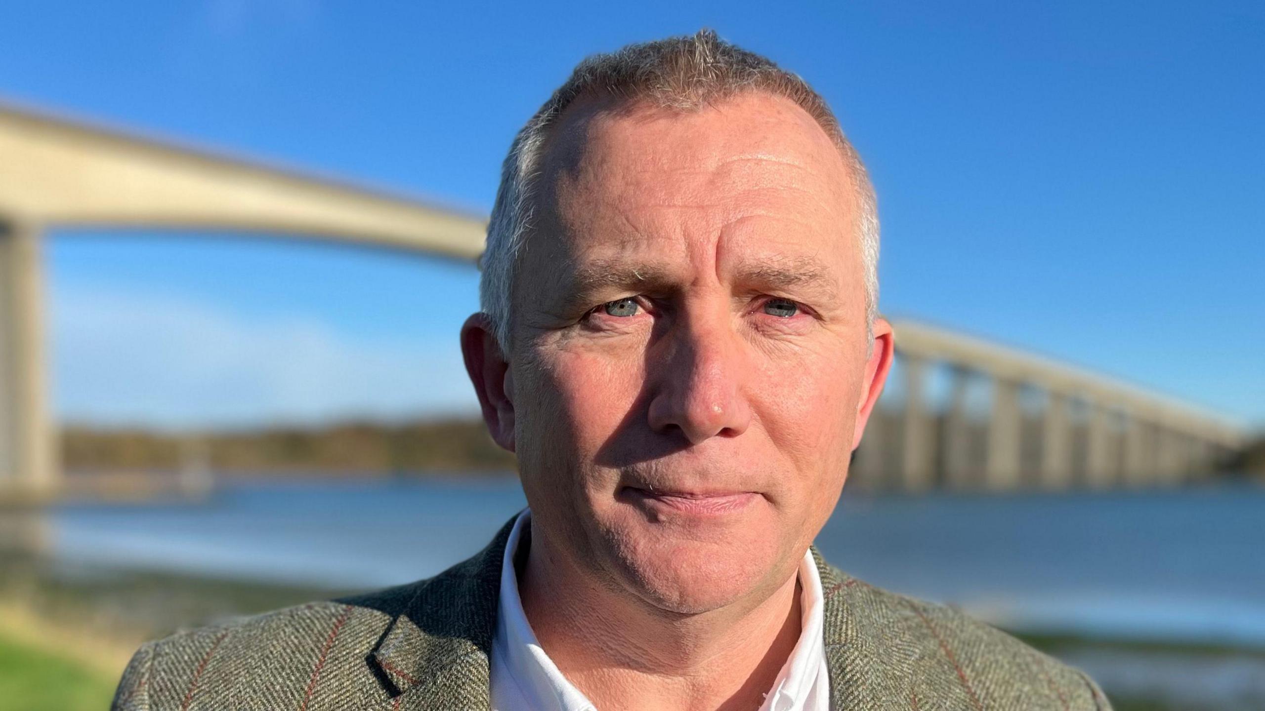 Mark Ling wearing a green jacket and a white shirt standing in front of the Orwell Bridge