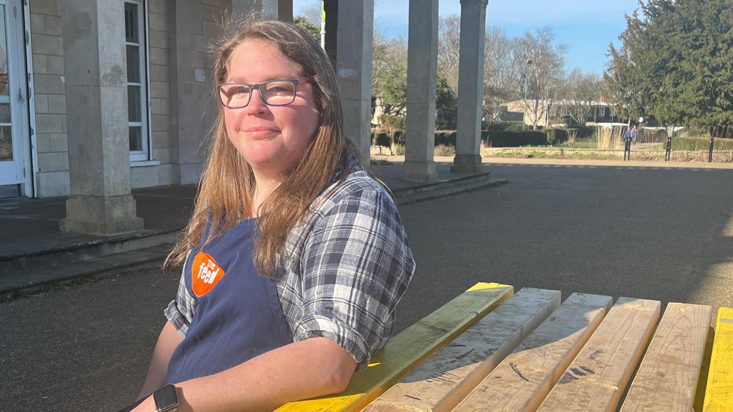 A woman with long brunette hair sitting on a wooden bench outside The Feed cafe. She is wearing a blue apron which says "The Feed" in white writing on an orange circle. She is wearing a blue checked shirt and black-rimmed glasses. She is looking at the camera and smiling.